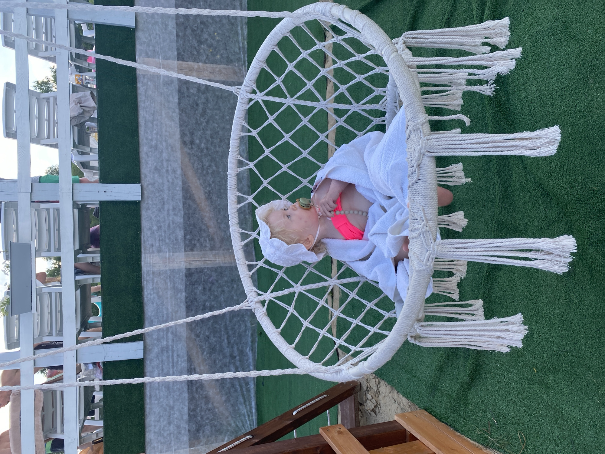 A young child is sitting in a white macrame hanging chair outdoors. The child, wearing a white bonnet and a neon pink swimsuit, is wrapped in a white towel. They are holding a pacifier with a beaded string. The chair is suspended by thick white ropes and has decorative tassels hanging from its bottom edge. The chair sits on a patch of artificial green turf, with sand visible to the left of the frame. In the background, a white wooden structure with rows of empty white lounge chairs is visible, suggesting an outdoor seating area, possibly a resort or beachside location. The sky is overcast. The location is identified as Колокша, Russia, though this information is not visually verifiable from the image alone.