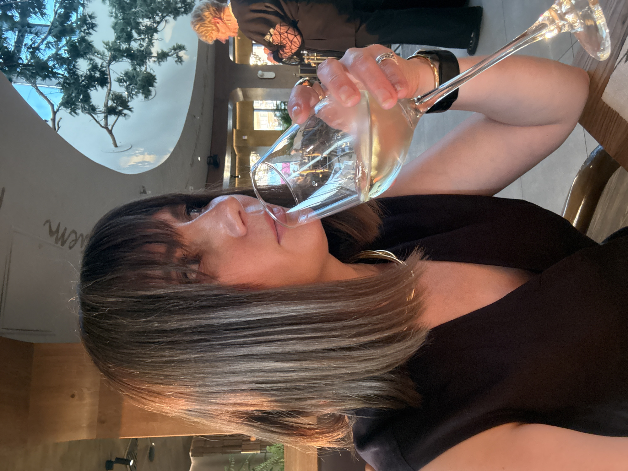 The image shows a medium shot of a woman with shoulder-length brown hair and bangs, wearing a black sleeveless top and a gold necklace. She is holding a glass of white wine to her lips, with her right hand supporting the stem of the glass. She is wearing several rings on her fingers and a black watch on her left wrist. The background is blurred but appears to be an indoor setting with wooden pillars, a curved ceiling, and a tree branch visible against a bright sky. Another woman with blonde hair is visible in the background, wearing a black top with lace detailing. The lighting suggests it is either daytime or a well-lit interior. There is no discernible information in the image to indicate the location is in the city of Izhevsk, Russia.