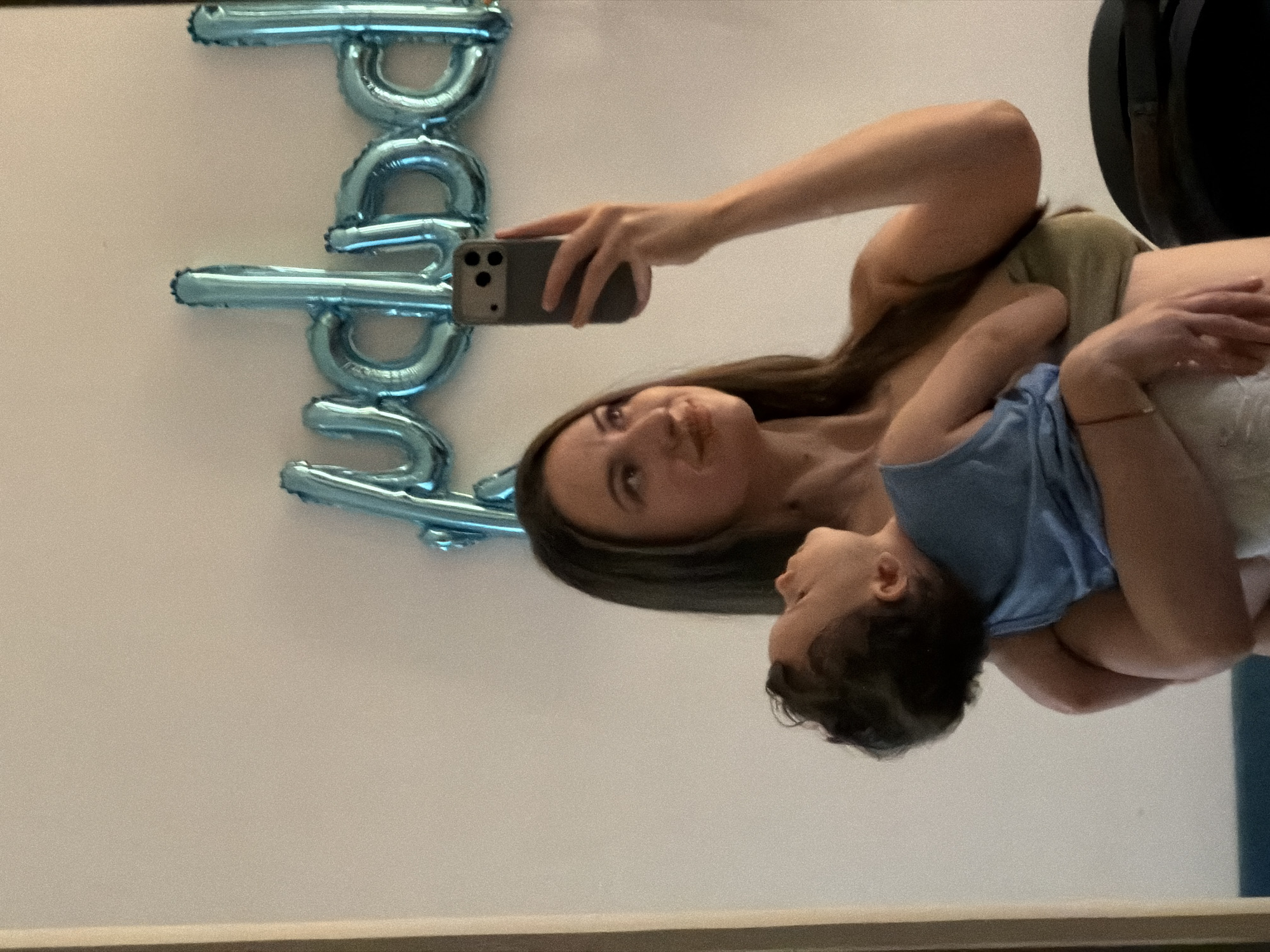 A woman is taking a mirror selfie while holding a baby. The woman has long, dark hair and is wearing a strapless top. She is holding the baby in her arms. The baby is wearing a blue tank top and is looking up. In the background, there are blue balloons that spell out "baby." The scene appears to be in a hotel room in Sharm El Sheikh, Egypt.