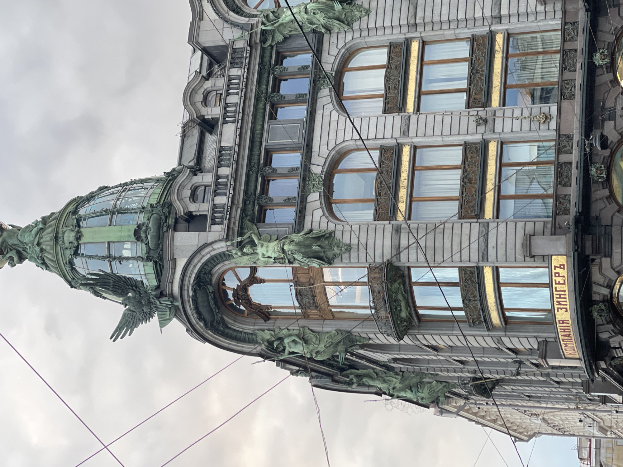 The image depicts a building facade in Saint Petersburg, Russia. The building is ornate, with a gray stone exterior, large arched windows, and decorative bronze or copper statues and adornments. A prominent dome with a bronze eagle figure is visible on the corner of the building. The windows have decorative frames and are partially covered with curtains. A sign with Russian text, likely identifying the building or a business within it, is on the side of the building. Overhead cables are visible in the foreground. The sky is overcast.