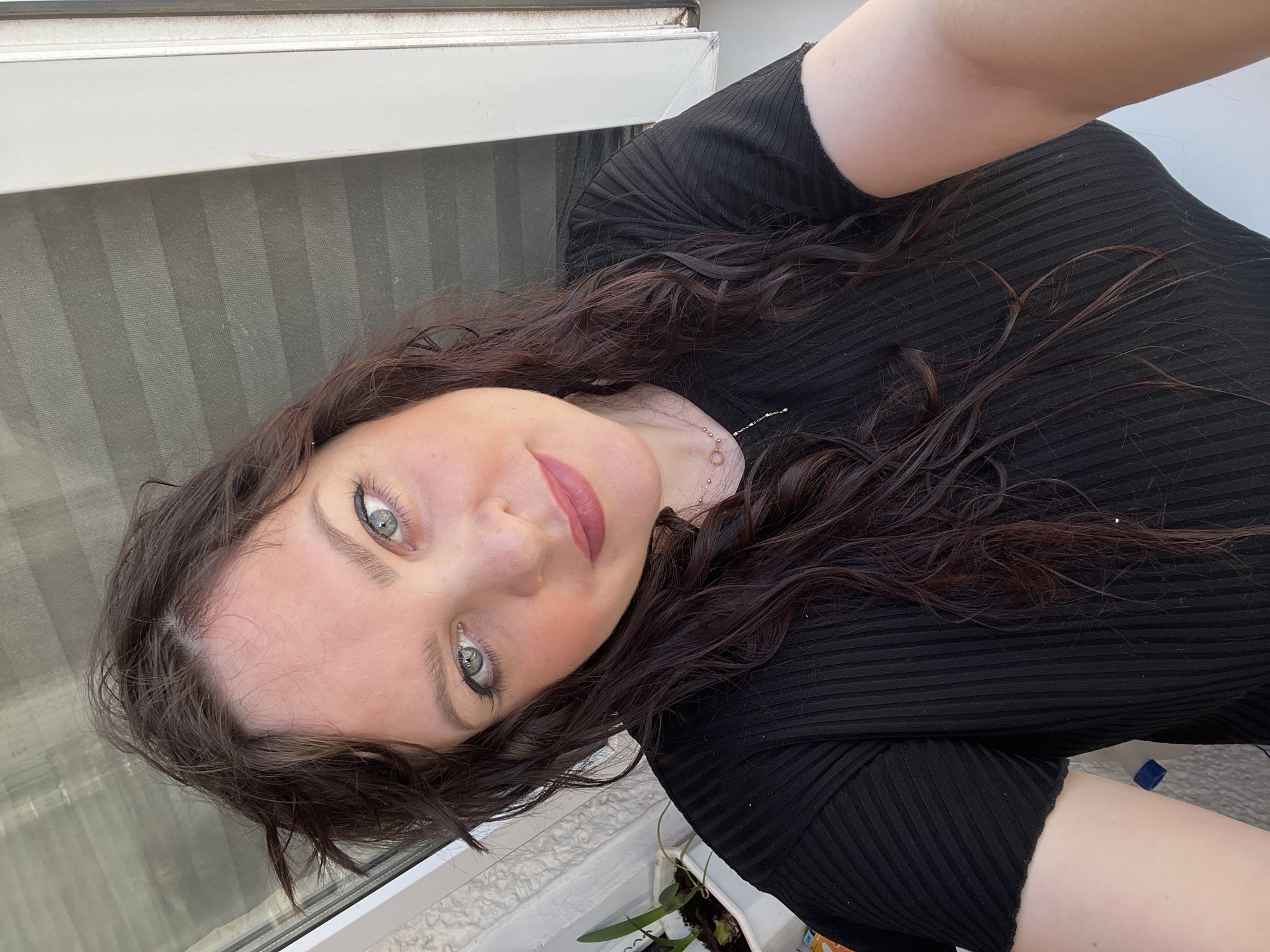 The image displays a close-up selfie of an adult female, located in the city of Noviy, Russia. The subject has dark brown, wavy hair, blue-grey eyes with dark eyeliner, defined eyebrows, and pink lipstick. She wears a black, short-sleeved, ribbed top and a thin gold-colored necklace featuring a circular pendant and interconnected circular elements. She is looking directly into the camera with a neutral expression. To her left, a window with horizontal blinds or a screen is visible through the glass, bordered by a white frame. To her right, a portion of a white textured wall or exterior surface is visible, alongside a white planter containing a green plant on what appears to be a windowsill or ledge. Natural light illuminates the scene from the right.