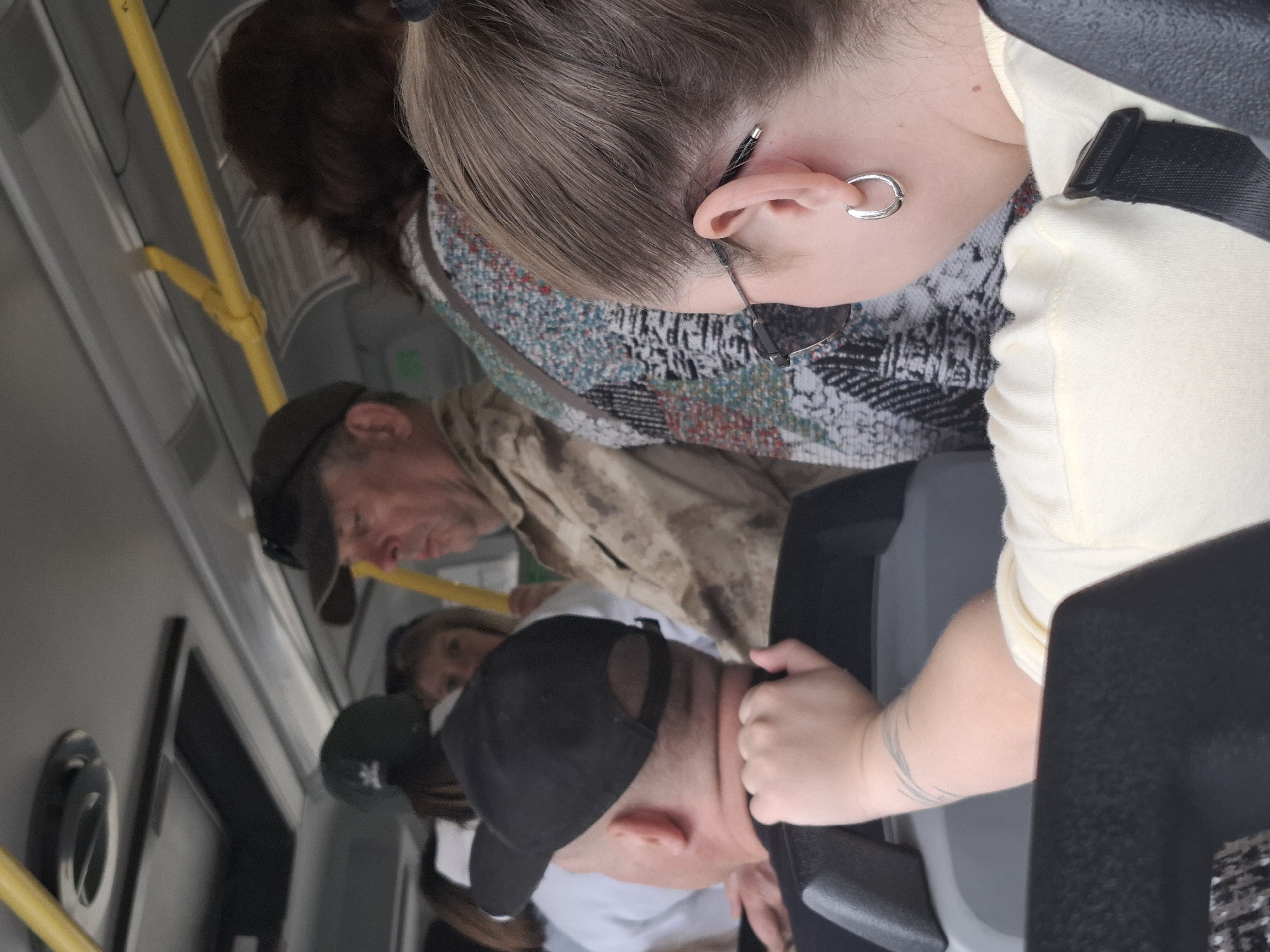 The image depicts a crowded public transportation setting, likely a bus, with multiple people visible. In the foreground, a person with short brown hair is wearing a light-colored shirt and a patterned sweater. The person is wearing glasses and a hoop earring. Adjacent to this individual is another person in a black baseball cap, appearing to be holding onto a seat. Behind them is an older man in a green cap and tan jacket. Further back, the presence of other individuals wearing various hats is noticeable. Yellow hand rails are visible in the background. The location is Chaikovsky, Russia.
