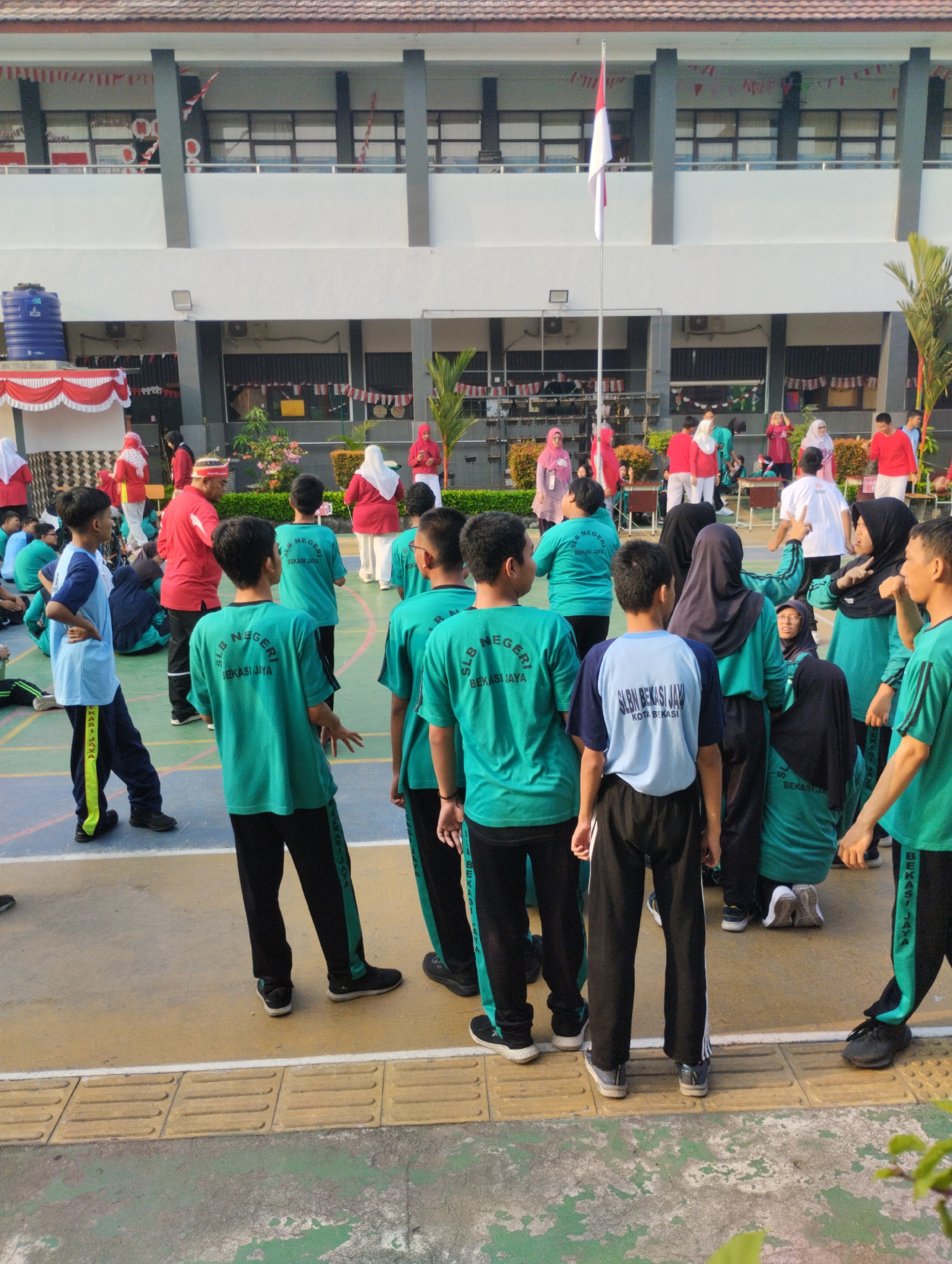 This image depicts an outdoor scene at a school in Bekasi, Indonesia. The foreground features a group of young people wearing teal t-shirts with text that appears to be "SLB NEGERI BEKASI JAYA". They are standing on a yellow and green court, with some in the process of walking or interacting. In the middle ground, there are more individuals in various colored clothing, including red and white, suggesting some form of event or activity. Behind them, a white building with multiple levels is visible, decorated with red and white banners. A flagpole with an Indonesian flag is also prominently displayed. In the background, palm trees and other greenery add to the setting. The overall impression is of a school gathering or sports event taking place in a sunlit environment.