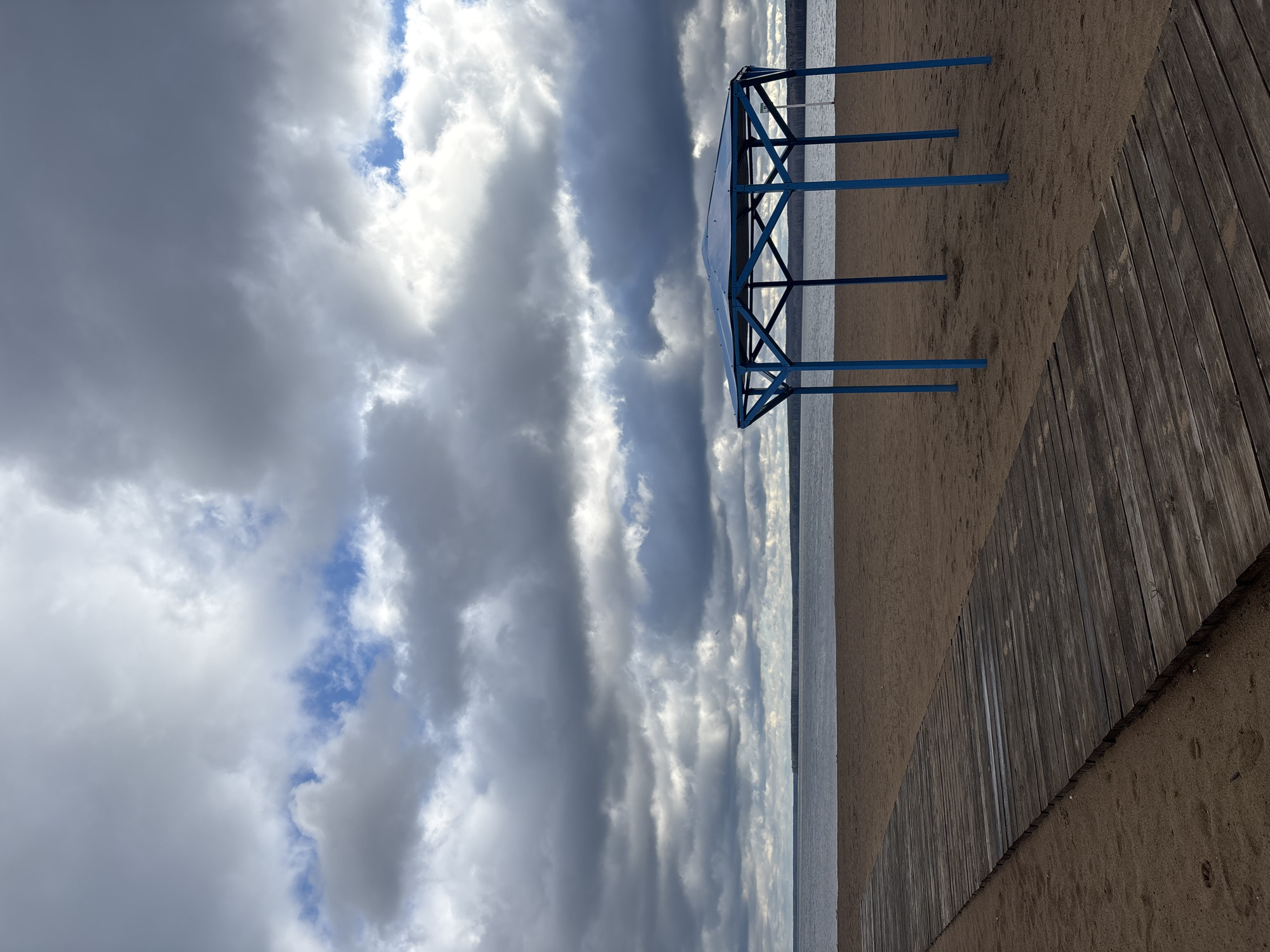 The image depicts a serene beach scene in Chaikovsky, Russia, under a dramatic sky. A large expanse of sandy beach stretches across the mid-ground, leading to a calm body of water, likely a lake or wide river, on the left side of the frame. On the right, a weathered wooden boardwalk runs diagonally across the sand, providing a path.
A prominent feature on the sandy beach is a blue metal frame structure, possibly a dismantled or unfinished canopy or small shelter, positioned closer to the water. Its skeletal form is open, without any covering, showcasing its crisscrossing supports.
The sky is a significant element of the photograph, dominated by a mix of heavy grey and brilliant white clouds. Patches of bright blue sky are visible in some areas, particularly peeking through breaks in the clouds, suggesting the sun is intermittently breaking through. The light creates varying tones on the clouds, from deep shadows to luminous highlights.
The scene appears devoid of people, contributing to a tranquil and possibly slightly melancholic atmosphere. There is no visible activity or event taking place. The overall impression is one of natural beauty with a touch of man-made presence, perhaps off-season or during a quiet period. No visible text is present in the image.