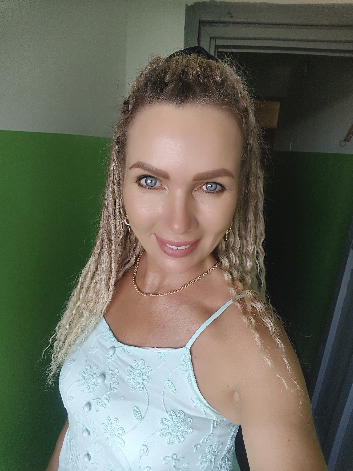 A medium shot, waist-up portrait captures a woman with blonde, curly hair styled in braids and a bun, facing forward. She has blue eyes, fair skin, and is wearing a light blue dress with floral embroidery and a gold chain necklace. Her smile is broad, revealing white teeth. The background features a two-toned wall, with the top section painted a light gray and the bottom section a vibrant green. To the right, a dark gray doorframe and a portion of a dark gray door are visible. The lighting is even, suggesting an indoor setting. The woman is positioned in the foreground, with the background slightly out of focus.