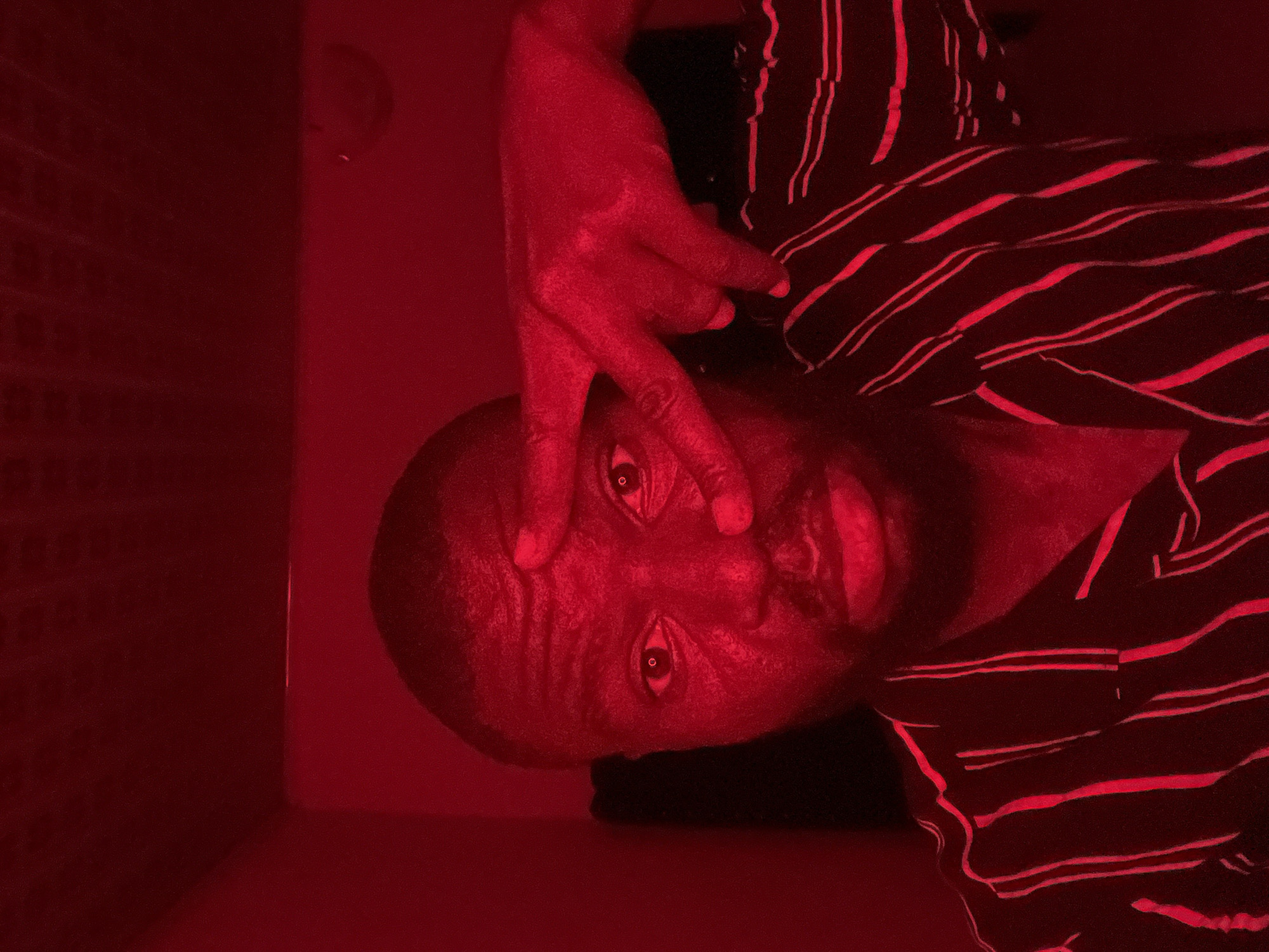 An intensely lit portrait features a person, likely male, with dark skin and a beard, looking directly at the camera. The entire scene is bathed in a strong, monochromatic deep red light. The person's head is positioned on the left side of the frame, with their upper body and a striped garment extending to the right. Their right hand is raised, with the index and middle fingers extended in a 'V' shape, held close to their right eye. The person's eyes are wide open, fixed on the viewer, contributing to the image's intensity. They are wearing a top or shirt with horizontal red stripes against a darker fabric, possibly black or dark blue. The background behind the person consists of a textured, possibly paneled, dark red surface on the left and a smoother, solid dark red wall on the right. The artificial red lighting creates a dramatic and enigmatic atmosphere, making it difficult to discern the exact details of the indoor setting or time of day. No text is visible.