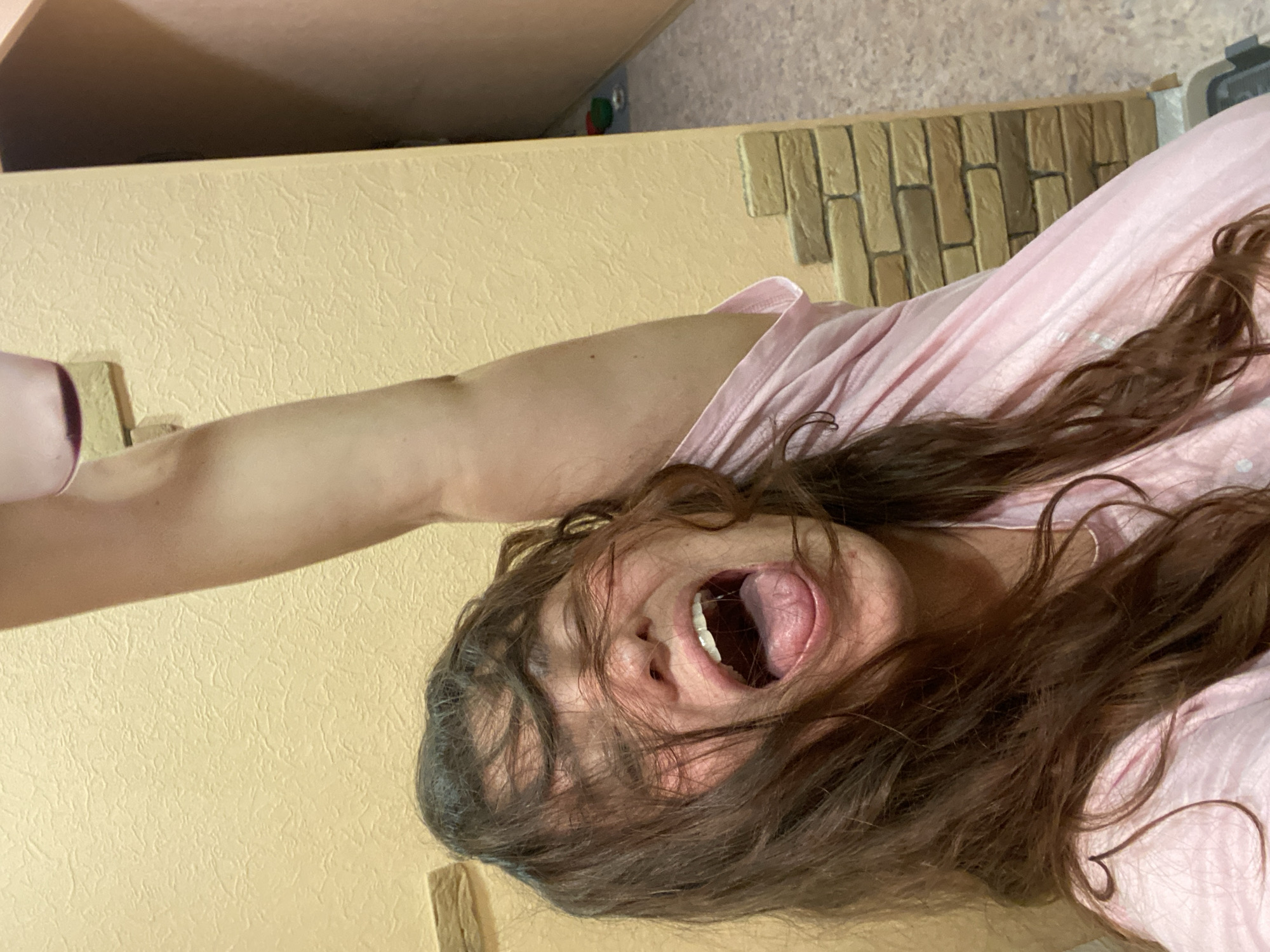 A woman with long, dark, wavy hair is captured in a close-up, high-angle shot, her face contorted in an open-mouthed scream or yawn. Her eyes are closed, and her hair obscures much of her face. She is wearing a pink top with a V-neckline. Her right arm is raised above her head. The background consists of a textured, pale yellow wall with decorative stone accents, suggesting an interior setting. The image does not provide any specific details that would allow for the identification of the city of Perm, Russia.