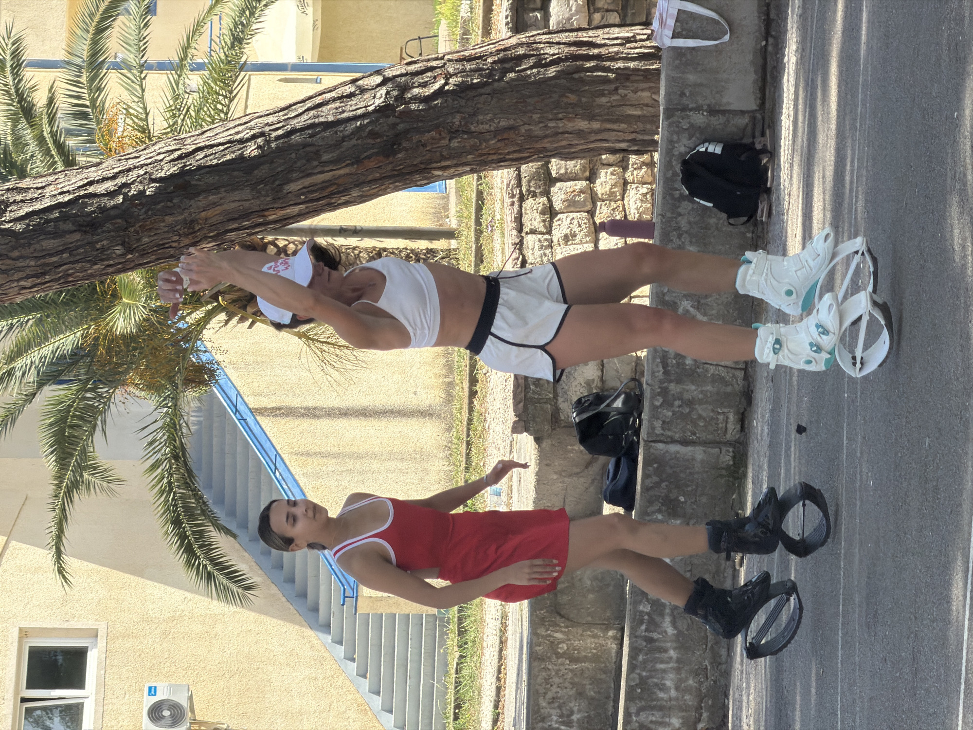 Two women are standing on a sidewalk on a sunny day. The first woman is closer to the viewer and is wearing a red dress and black spring-loaded boots. The second woman is further from the viewer and is wearing a white sports bra, white shorts with black trim, a white visor, and white spring-loaded boots with blue accents. The woman in the white outfit is stretching her arms. A tree trunk is above the second woman. Behind the first woman is a set of stairs, a building with an air conditioning unit, and a palm tree. A black backpack is on the ground behind the second woman. The location is Budva, Montenegro.