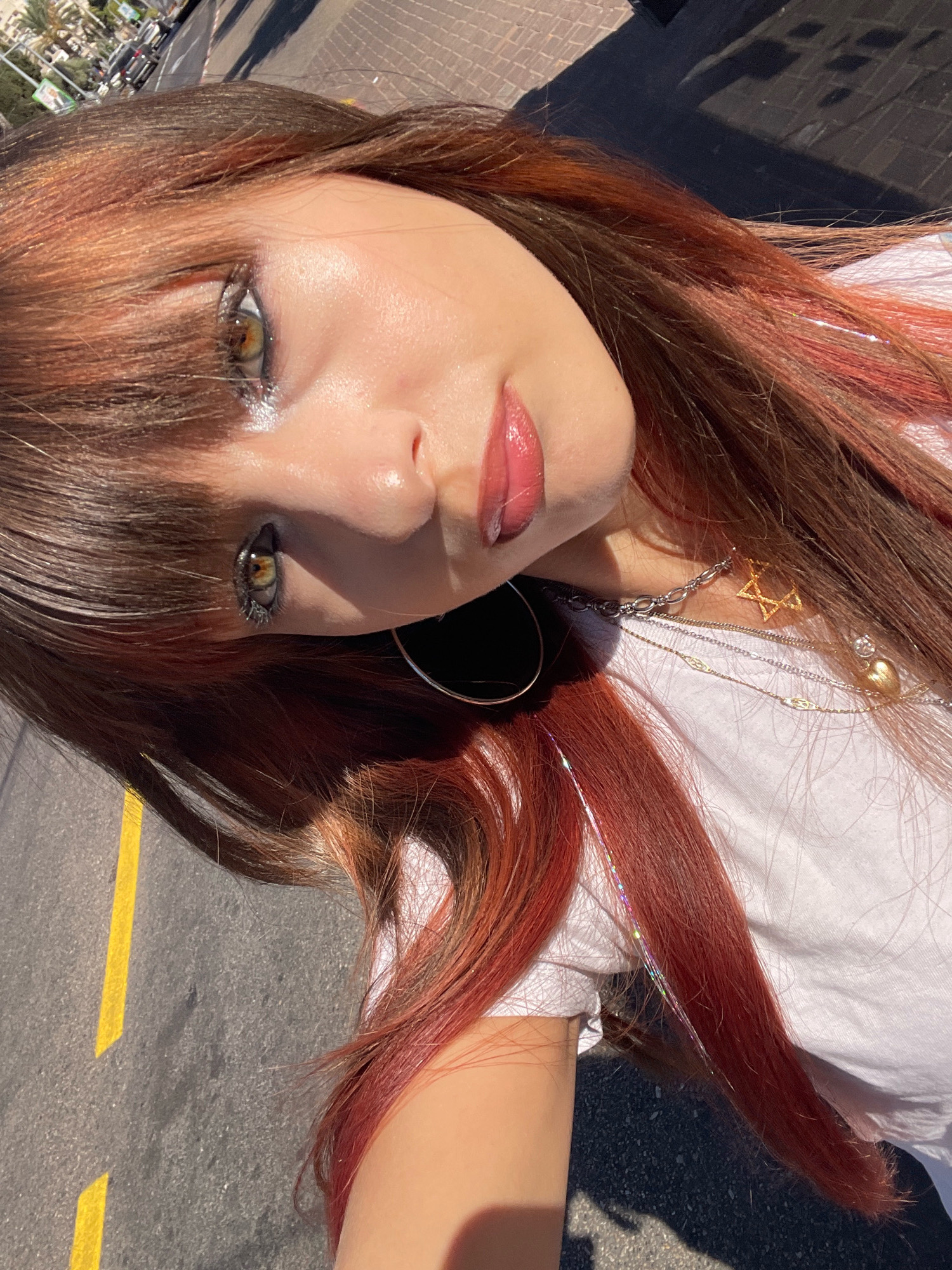 The image is a close-up, high-angle selfie of a young woman with auburn hair, accented with pink and iridescent streaks, taken outdoors on a sunny day. She is wearing a white t-shirt and several necklaces, including one with a Star of David pendant and another with a heart charm. Her makeup includes bold eyeliner and glossy pink lipstick. The background suggests an urban environment, possibly a street with a paved surface marked with yellow lines, and a glimpse of palm trees and buildings in the distance. The overall scene conveys a sense of modern, casual style.