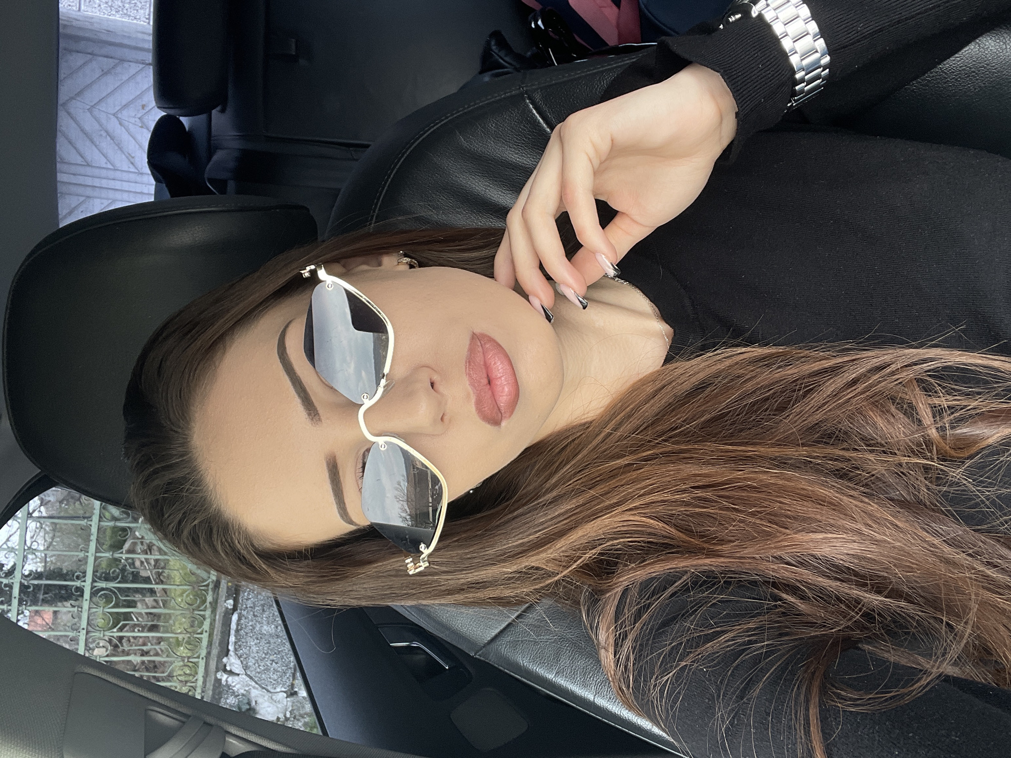 The image shows a woman lying in a car. She is wearing sunglasses and a black top, with a silver watch on her wrist. Her hair is brown. The location is likely inside a car in Tserova Koria, Bulgaria, indicated by details visible through the car window.