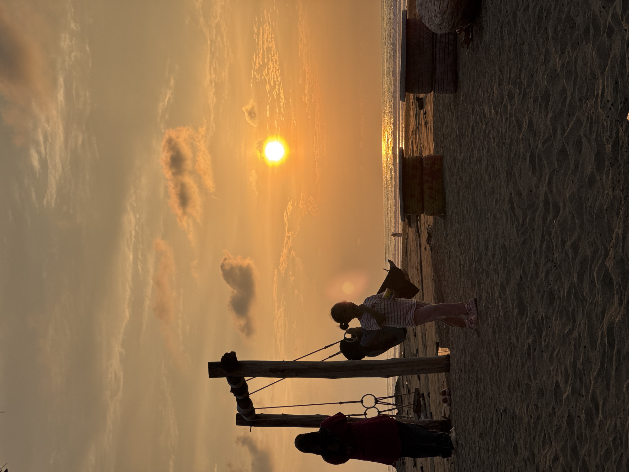 In Reudeuep, Indonesia, a beach scene unfolds at sunset. The sky is ablaze with a warm orange hue, the sun a bright orb hovering over the ocean horizon, its reflection shimmering on the water's surface. Two figures, silhouetted against the vibrant sky, stand on a sandy beach near a wooden swing set. One figure, likely an adult wearing a headscarf and a maroon top, stands facing away from the viewer. Beside them, a child in striped clothing plays near the swing. The beach is dotted with weathered tire tables, remnants of what might be a seaside gathering spot. The tide appears low, exposing wet sand and scattered debris. The atmosphere is serene, capturing a moment of tranquility as day transitions to night.