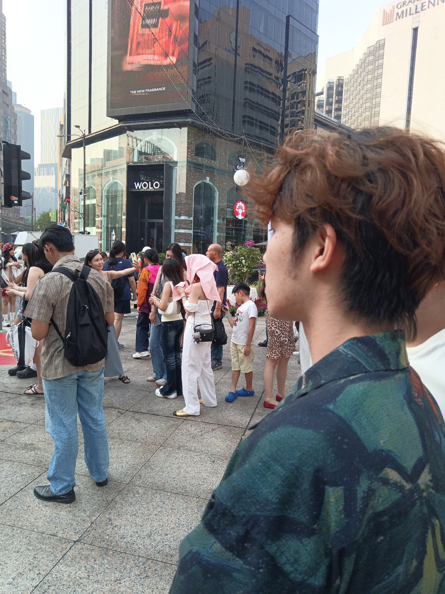 The image depicts a street scene in Kuala Lumpur, Malaysia, likely within a bustling commercial district. In the foreground, a young man with curly brown hair is seen from behind, wearing a dark green shirt with an abstract pattern. He is facing away from the camera, looking towards a group of people.
The middle ground features a diverse crowd of individuals. A man in a checkered shirt and jeans, with a backpack, stands facing the group. Among the others are women in casual attire, some holding phones, suggesting they are taking pictures or interacting with social media. One woman is holding a pink umbrella. A young boy in a white t-shirt and shorts is also present.
The background is dominated by modern architecture. A large building with a glass facade displays an advertisement for "THE NEW FRAGRANCE." Below it, another building has "WOLO" prominently displayed. Reflections of city buildings are visible on the glass surfaces. Traffic lights and street lamps are also present, indicating a public urban space. The overall atmosphere appears to be that of a busy day in the city, with people engaged in various activities.