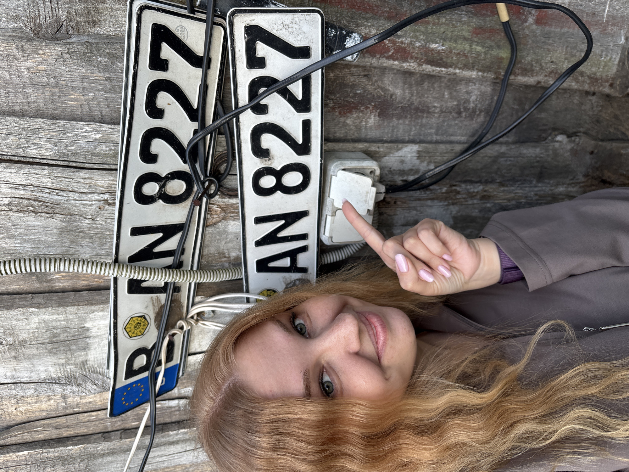 A fair-skinned female with long, wavy, light reddish-blonde hair, light green eyes, and a smiling expression is featured in the foreground. She wears a grey-brown jacket over a purple garment and has light pink nail polish. Her right index finger points towards a light grey electrical outlet unit with two sockets, mounted on a weathered wooden plank wall.
Above the outlet, two rectangular license plates are affixed to the wall, partially obscured by electrical wires. The top plate shows a blue European Union emblem with yellow stars, followed by "D", a circular yellow and black emblem, and then "N 8227". The bottom plate is fully visible, displaying "AN 8227". Both plates appear worn and dirty. Numerous black and white electrical wires, some twisted, run across the scene, and a vertical ribbed flexible conduit is visible on the right. The wooden background consists of aged, vertical planks, showing grey discoloration and natural wear, with a patch of faded red paint visible on the lower right. The scene is located in Rozhdestveno, Russia.