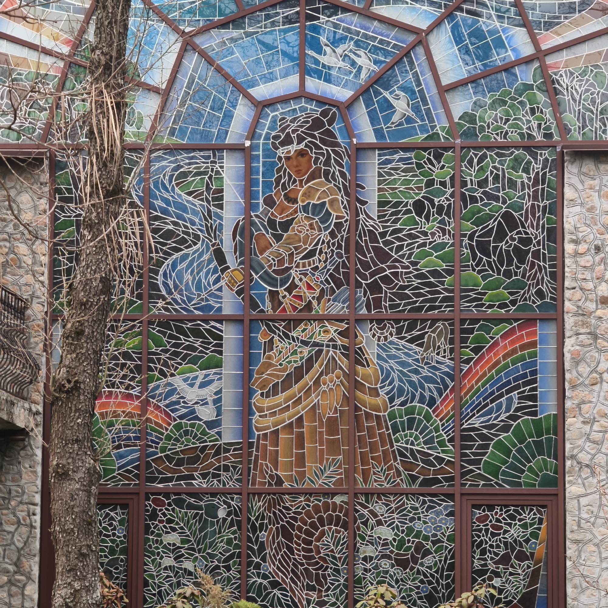 A detailed stained-glass window dominates the view, showcasing an indigenous woman holding a child, surrounded by natural elements such as trees, water, birds, and a rainbow. The window is sectioned with dark brown framing, and part of a tree trunk obscures the left portion of the artwork. The window is set into a stone wall with an attached wrought-iron balcony, located in Капцёўскі сельскі Савет, Belarus.