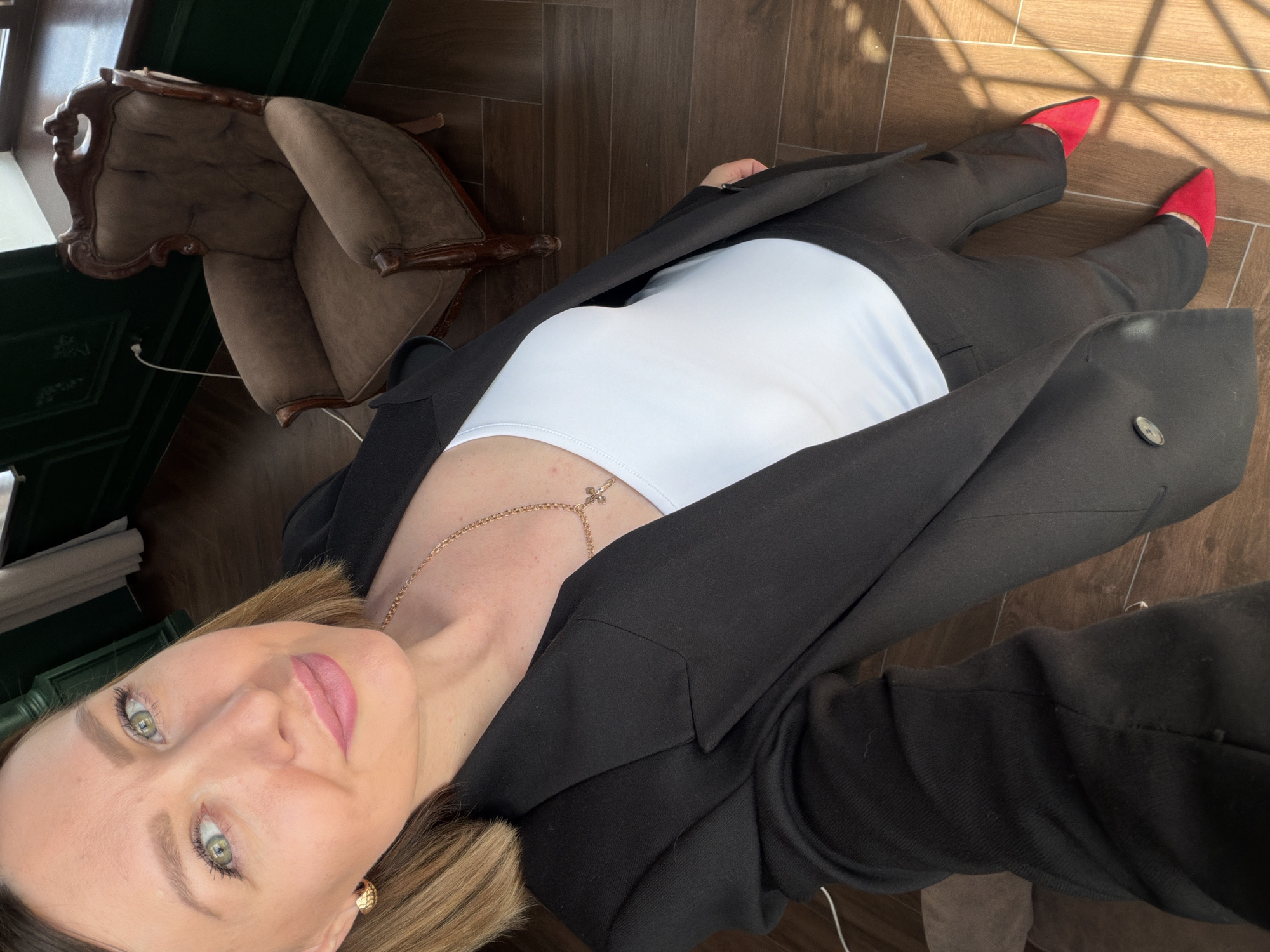 A woman in a black blazer, white top, and black pants is indoors, likely in Rostov-on-Don, Russia. She's wearing red shoes. A brown armchair is visible on the left side of the frame. The floor is brown and has a wood-like pattern. Natural light is present, creating shadows on the floor. The woman is looking at the camera with a slight smile. She's wearing a gold necklace and earrings.