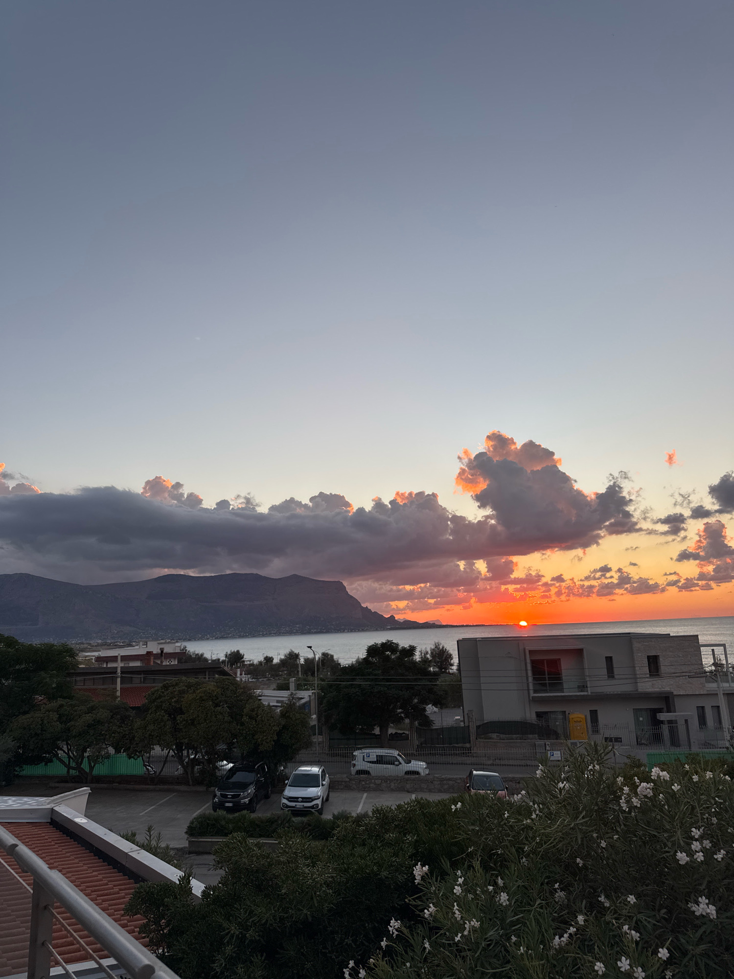 The image depicts a sunset over the sea in Capaci, Italy. The sun, a bright orange orb, is setting on the horizon, casting a warm glow across the sky and illuminating the clouds. The clouds are a mix of dark grays and vibrant oranges, creating a dramatic and colorful scene. In the foreground, there is a building with a modern facade, and below it, a parking lot with several cars. Trees and some white-flowered bushes are also visible. In the background, a mountain range can be seen across the water. The overall impression is one of a tranquil evening in a coastal town.