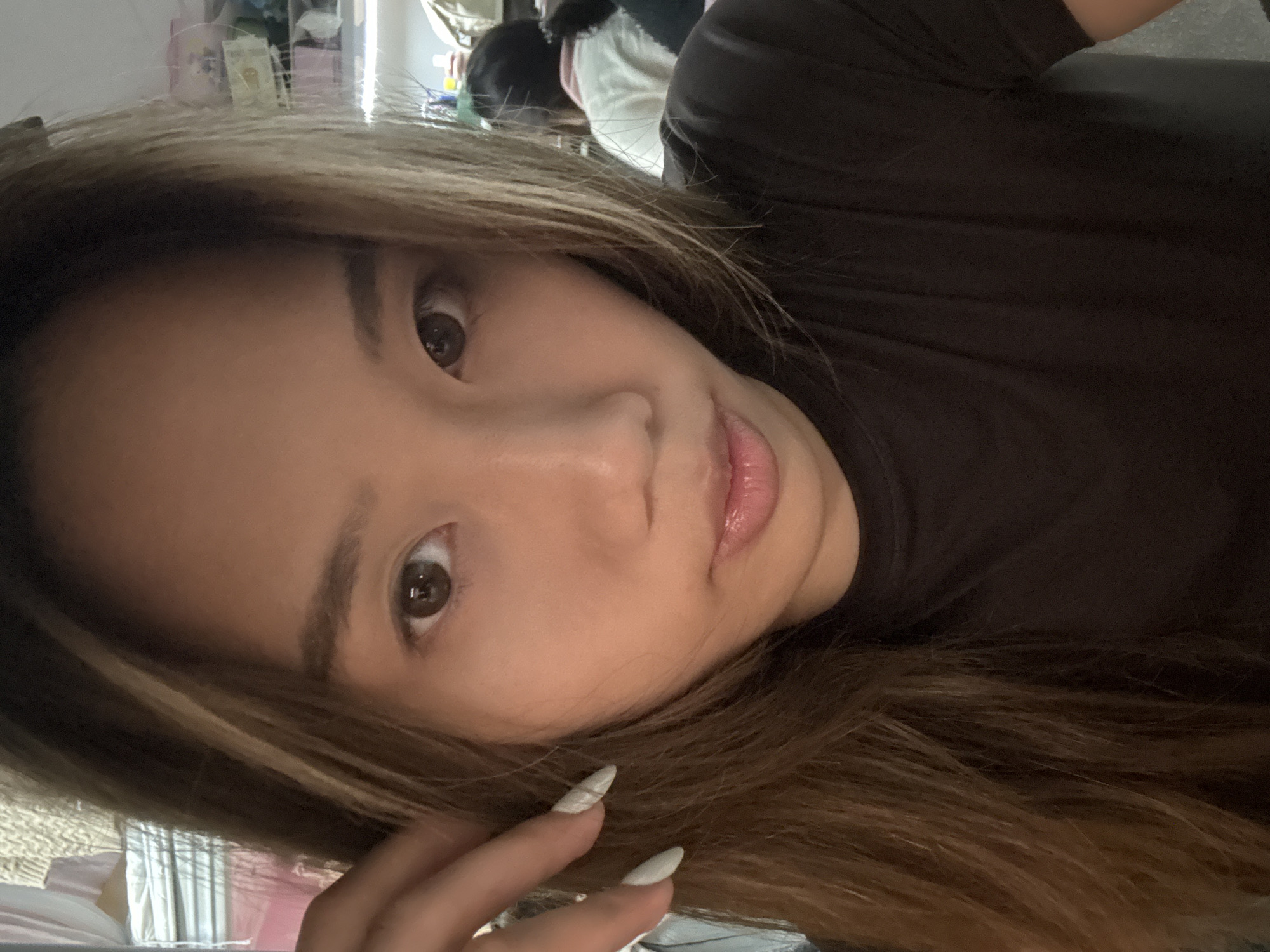 A close-up, angled selfie of a woman with long brown hair with blonde highlights. She is wearing a dark-colored top. Her hand with long, white, pointed fingernails is near her face. The background is blurry, but appears to contain clutter, possibly in an indoor setting. The location is not discernible based on the image.
