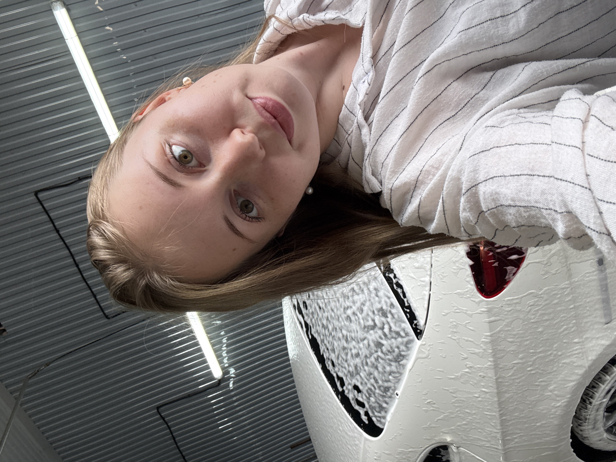 A young woman with blonde hair, wearing a white and black striped shirt, takes a selfie with a white car covered in foam in the background. The car's rear window and part of its side are visible, along with a red taillight. The setting appears to be a car wash, with corrugated metal walls and fluorescent lighting overhead. The woman is positioned in the foreground, with her face tilted towards the camera.