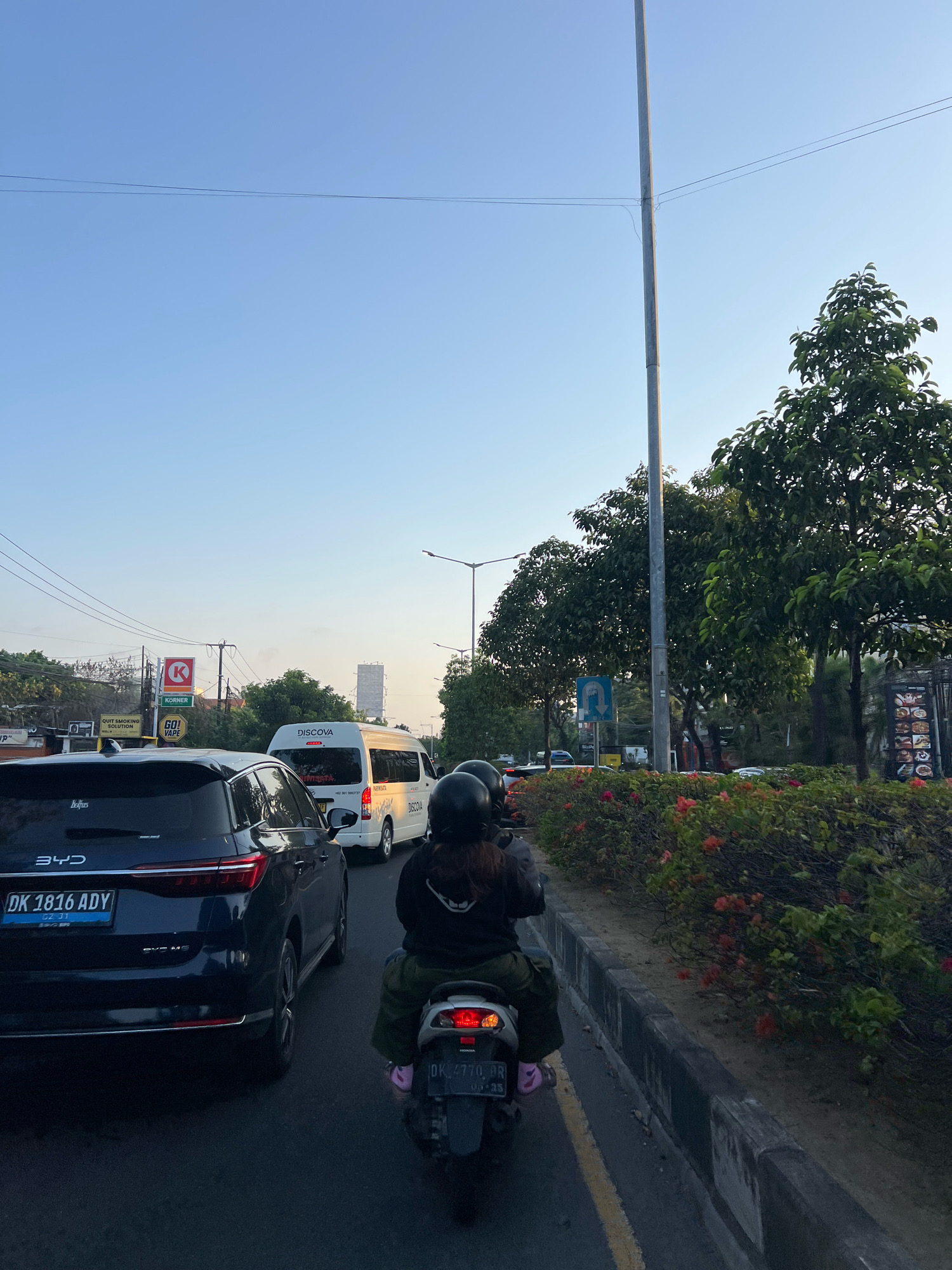 An eye-level, medium shot captures a traffic scene on a street in Kuta, Indonesia, as dusk settles. The sky is a clear, pale blue, transitioning to a lighter hue near the horizon. A tall, slender utility pole dominates the mid-ground, with wires extending horizontally across the frame. To the left, a dark blue BYD electric SUV with a license plate reading "DK 1816 ADY" is stopped in traffic. To the right, a motorcycle with two occupants is also stationary. The rider, wearing a black helmet and hoodie, is facing away from the camera, and a second passenger, also in a black helmet, is behind them. A white passenger van with "DISCOVA" branding is visible ahead of the SUV. The roadside is lined with lush green trees and flowering bushes. Billboards and signs, including one for "KORNER" and "GO! VAPE", are present on the left side of the street. The road surface is asphalt with yellow lane markings.