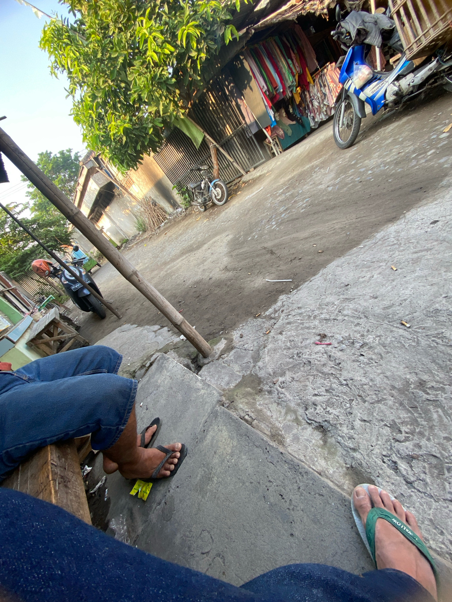 In Gurah, Indonesia, a low-angle, wide shot captures a dusty street scene on a sunny day. In the foreground, two individuals, partially visible from the waist down, are seated. One person, on the left, wears blue jeans and black flip-flops, with their legs crossed. A piece of yellow paper lies beneath their feet. The other person, on the right, also wears blue jeans and green flip-flops.
The environment consists of a dirt and concrete road, littered with cigarette butts. To the left of the road, a wooden structure is visible, supporting what appears to be a rudimentary stall with clothing hanging from it. Further down the street, a blue and white Suzuki motorcycle with a cargo rack is parked. Another dark-colored motorcycle with an orange helmet on it is also present. Overhead, a large tree with green leaves casts shadows, and a corrugated metal structure, possibly a building or roof, is visible behind the clothing stall. The overall impression is of a casual, everyday scene in a semi-urban or rural area of Indonesia.