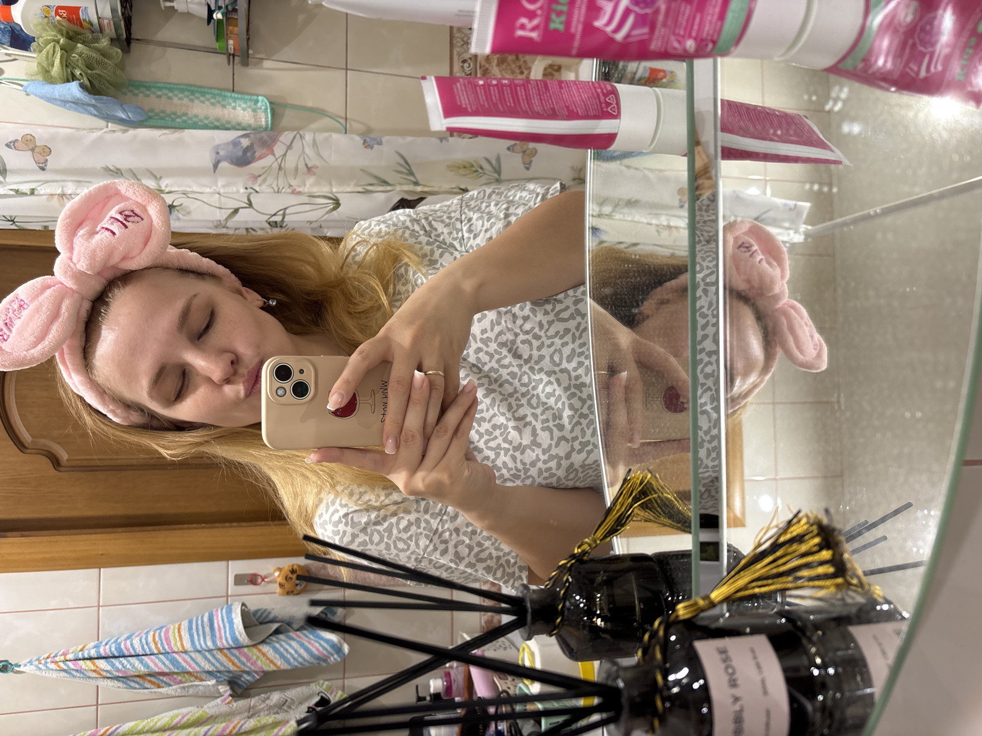 A fair-skinned woman with blonde hair and a pink bunny-ear headband is taking a selfie in a bathroom mirror. She is wearing a gray and white patterned top. The phone case has a picture of a wine glass on it. The background includes a shower curtain with a bird and butterfly pattern, bath products, and a black bottle with reeds and a yellow tassel. The bottle has the words "Bubbly Rose" on it. A towel is also hanging. The location is not specified as Rusanovo, Russia.