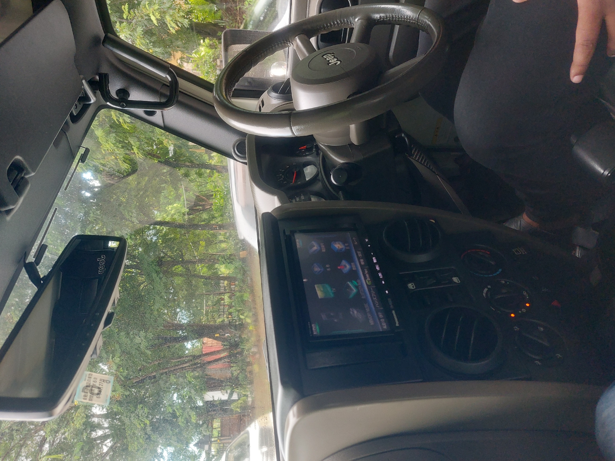 The image shows the interior of a Jeep vehicle, focusing on the dashboard, steering wheel, and rearview mirror. The Jeep logo is visible on the steering wheel. The dashboard features a screen displaying various icons and controls for air conditioning. The rearview mirror is present.
Outside the vehicle, through the windshield, trees and some buildings are visible. There is a sticker on the windshield with Japanese text. Based on the windshield sticker, the car might be in Japan. Dar es-Salaam, Tanzania cannot be confirmed as the location. There are no people prominently visible in the image except a person partially shown on the right side.