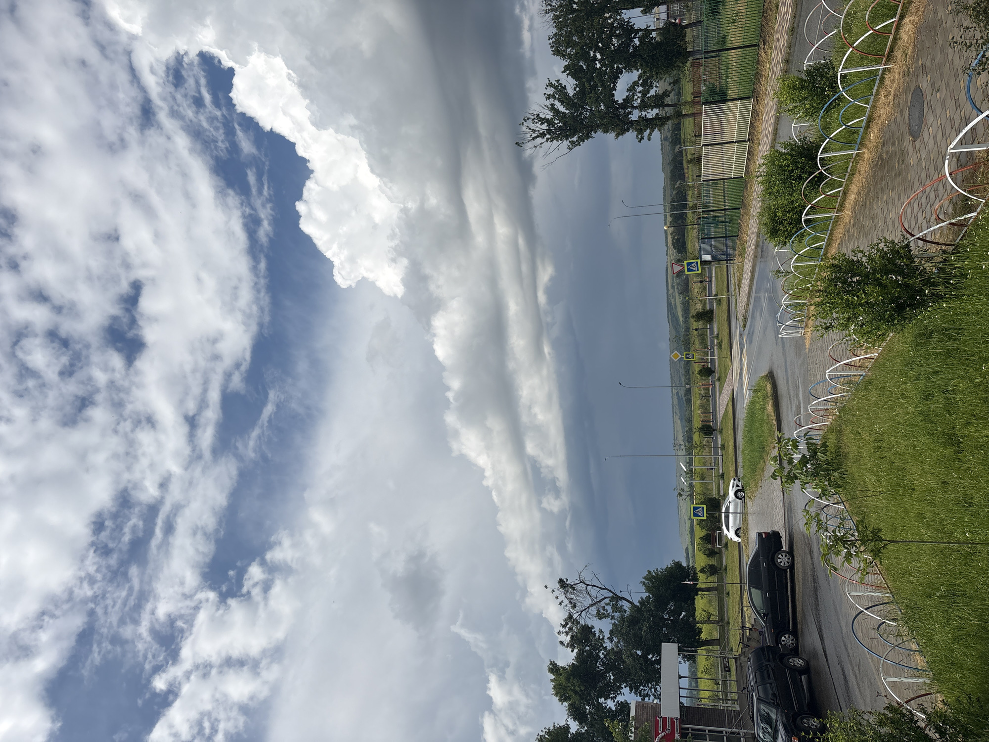 In Mikhaylovsk, Russia, a scene unfolds under a dramatic sky. The weather appears to be changing, with large, dark storm clouds gathering on the horizon, contrasting with brighter, white clouds in the foreground. The atmosphere is likely humid and potentially cool.
The image captures a street intersection, with wet asphalt suggesting recent or ongoing rain. A black SUV is parked on the left side of the road, followed by a black sedan. Further down the road, a white sedan is visible, approaching the intersection. Traffic signs are present, including yield signs and a pedestrian crossing warning sign.
A low, colorful fence with arched supports, painted in red, white, and blue, runs along the edge of a paved walkway and a grassy area. This fence delineates a pathway from a well-maintained grassy verge, which is dotted with small white flowers. Trees, some bare and others with full foliage, stand to the left and right of the frame. In the background, a green, hilly landscape stretches towards the horizon, partially obscured by the clouds. A metal fence runs along the right side of the image, behind which is a field. A building with a red awning is partially visible on the far left.