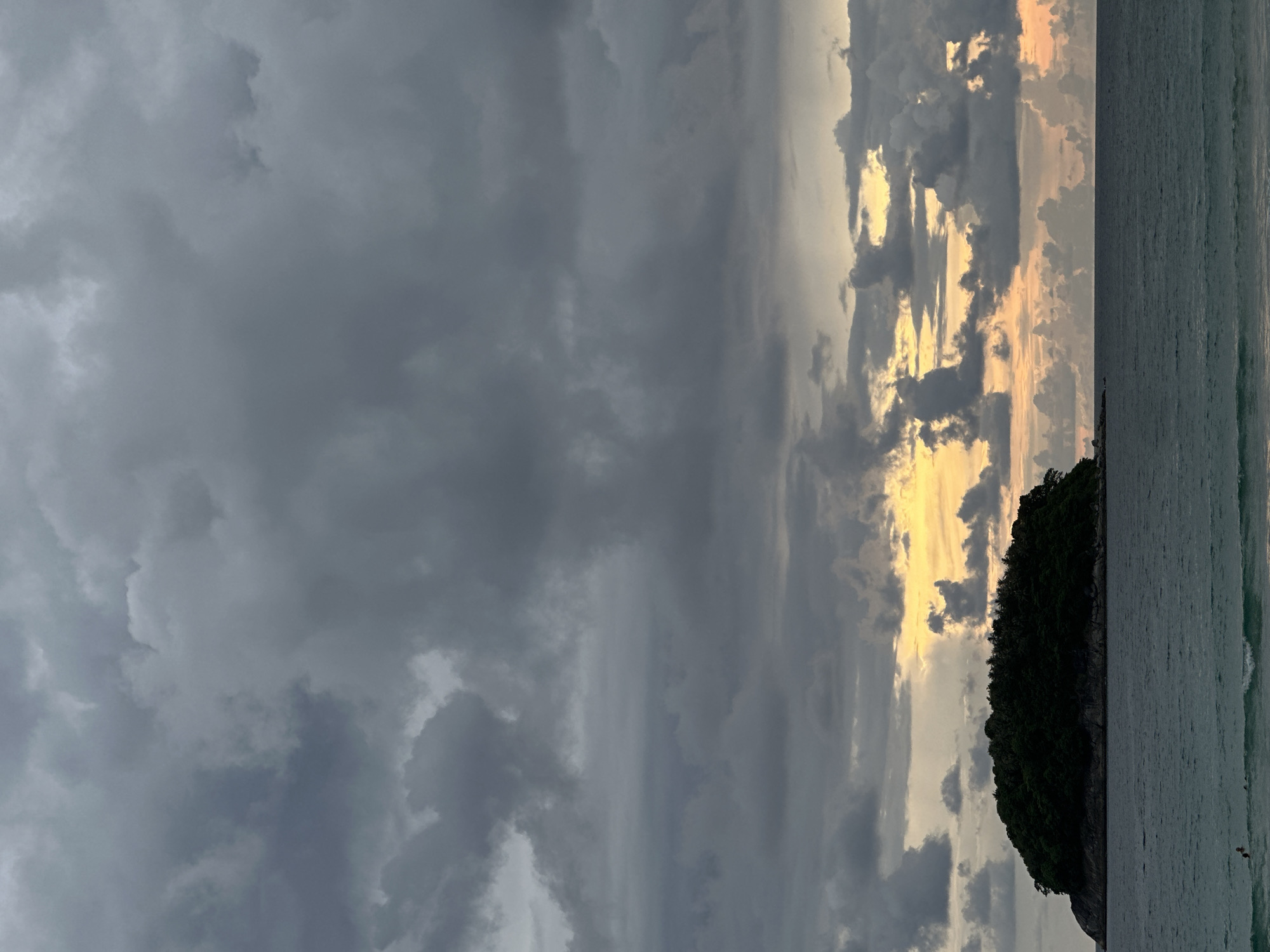 The image depicts a cloudy sky over the ocean, with a small, tree-covered island in the distance. The sky is filled with dark gray clouds, with a hint of yellow and orange light peeking through on the horizon, suggesting either sunrise or sunset. The ocean is a deep blue-green, with gentle waves visible. The island is a dark silhouette against the sky, with dense foliage covering its entirety. Two small figures are visible in the water in the lower left corner of the image, appearing to be swimming. The scene suggests a natural, tranquil environment, likely a coastal area. The location is Karon, Thailand.