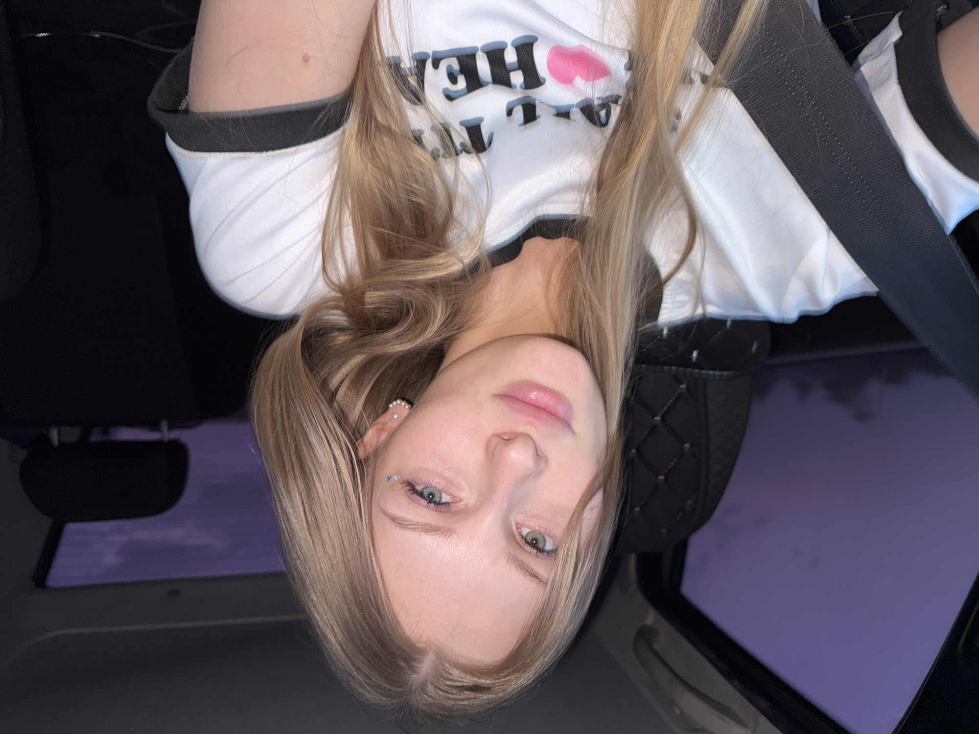 A young woman with blonde hair and light-colored eyes is pictured inside a car during twilight. She is wearing a white t-shirt with black trim and text that includes the word "HEART" and a pink lipstick imprint. A seatbelt is visible across her chest. The background shows the interior of the car and a purple twilight sky visible through the window. The scene is indoors, within a vehicle, and does not provide any specific information about the city of Perm, Russia.