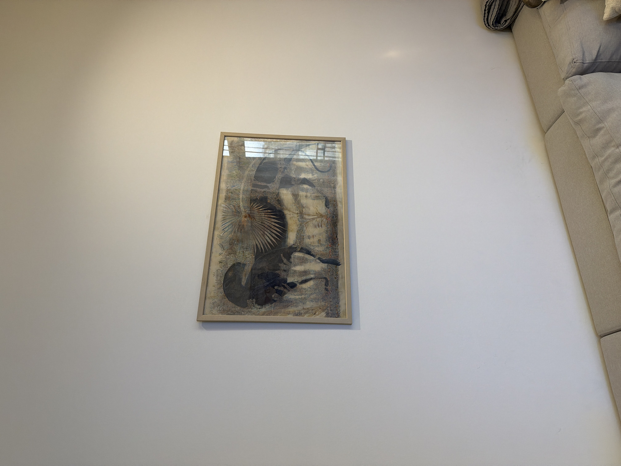 The image shows a framed piece of artwork hanging on a white wall. The artwork is abstract and features a mix of colors, including blues, browns, and creams, with a central shape that could be interpreted as an animal or creature. The artwork is rectangular in shape and framed with a thin, light-colored frame. A section of a light beige couch is visible on the right side of the image and is positioned close to the white wall. The wall and the setting suggest an indoor environment. Without additional context, specific location information about Tbilisi, Georgia, is impossible to verify.