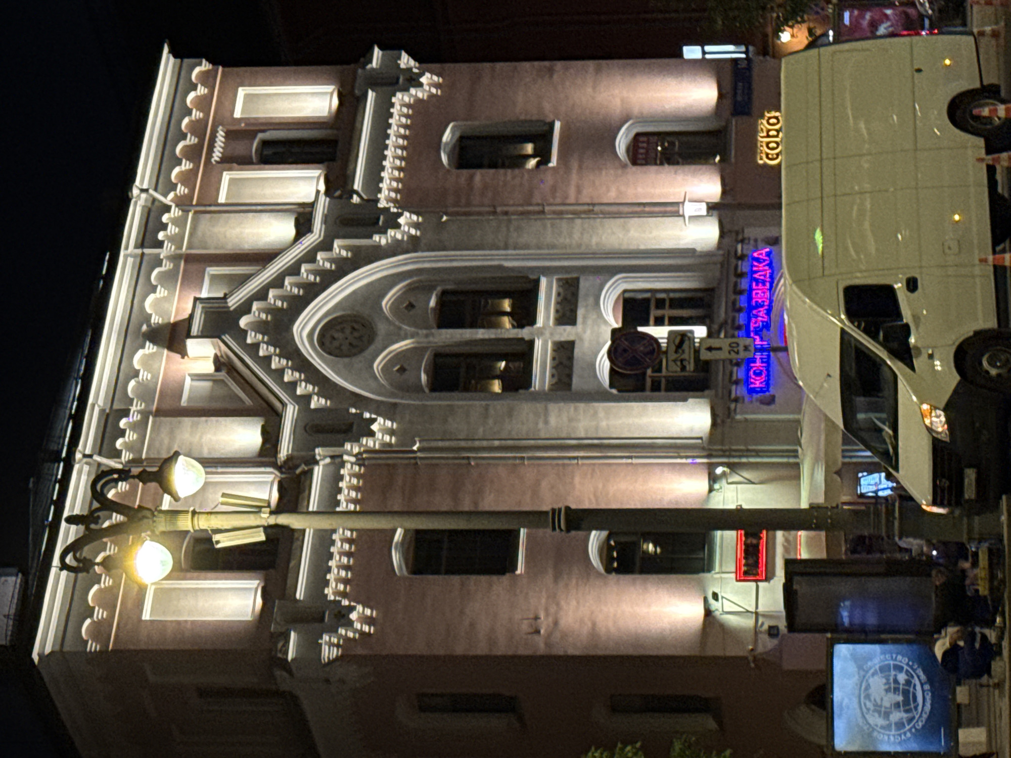 The image shows a building at night in Moscow, Russia. The building has a decorative facade and windows, and is illuminated by streetlights. A white van is parked in front of the building. There are signs and street furniture, including a lamppost, visible in the foreground. The sky is dark and the environment appears urban. A blue neon sign says "Конкурс Разведка" in Russian. The sign above reads "Собака". People are standing by the van.