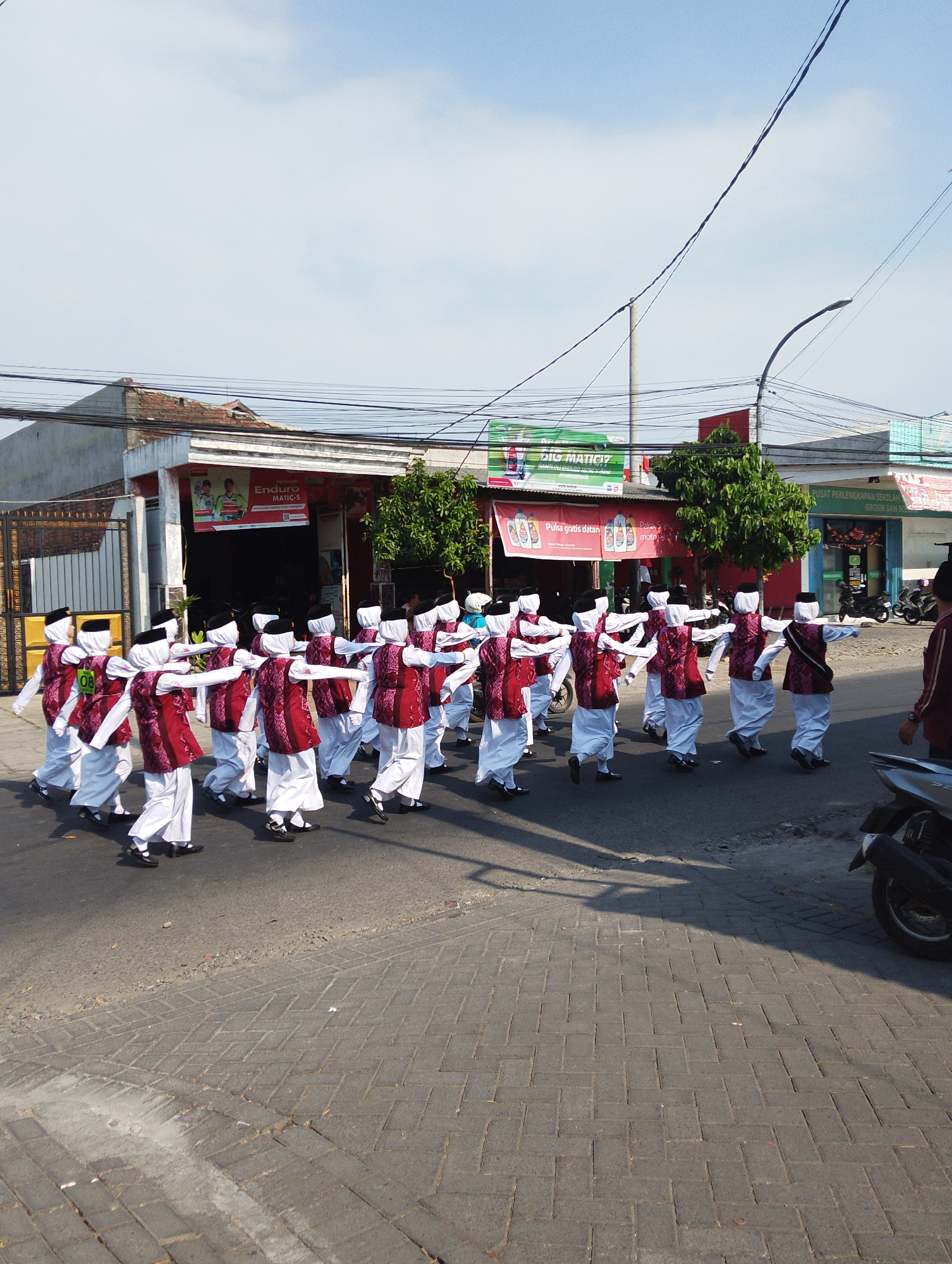 In Manaruwi, Indonesia, a group of approximately 20 children, dressed in matching white outfits with red patterned vests and black head coverings, march in formation along a paved street. They are performing some form of synchronized movement, with their arms extended to the sides. The street is lined with buildings, including a store with a red awning and signs advertising "Enduro MATIC-S" and "PULSA GRATIS DATAN MOTOR." Another building to the right appears to be a school supply store. Overhead, power lines crisscross the sky, and a street light is visible in the distance. A parked motorcycle is on the right side of the street. The overall scene depicts an outdoor event or parade on a bright, sunny day.