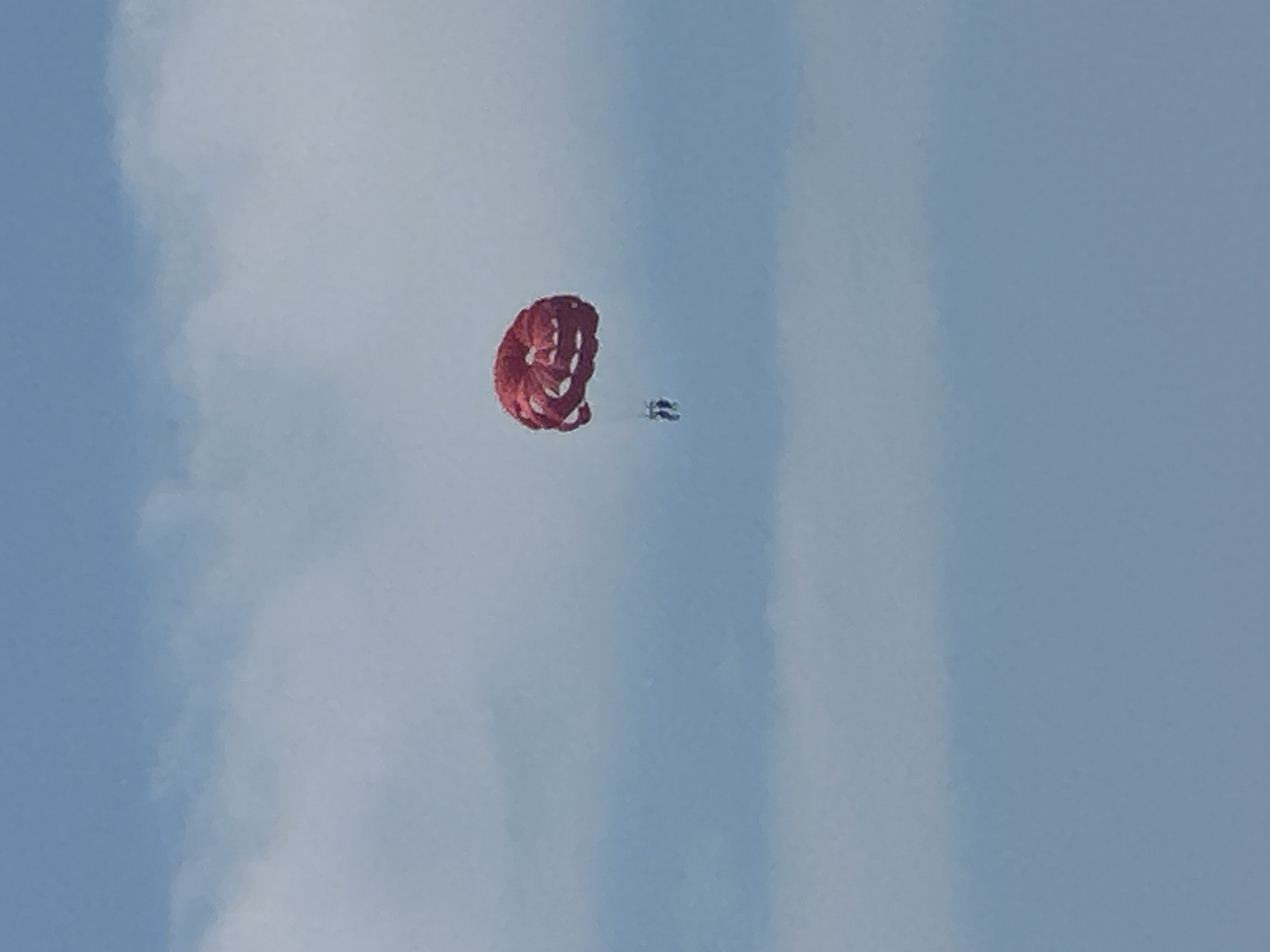Two people are parasailing in the sky over Budva, Montenegro. The red parachute is visible against a clear blue sky with some white clouds. The parasailers are suspended below the parachute.