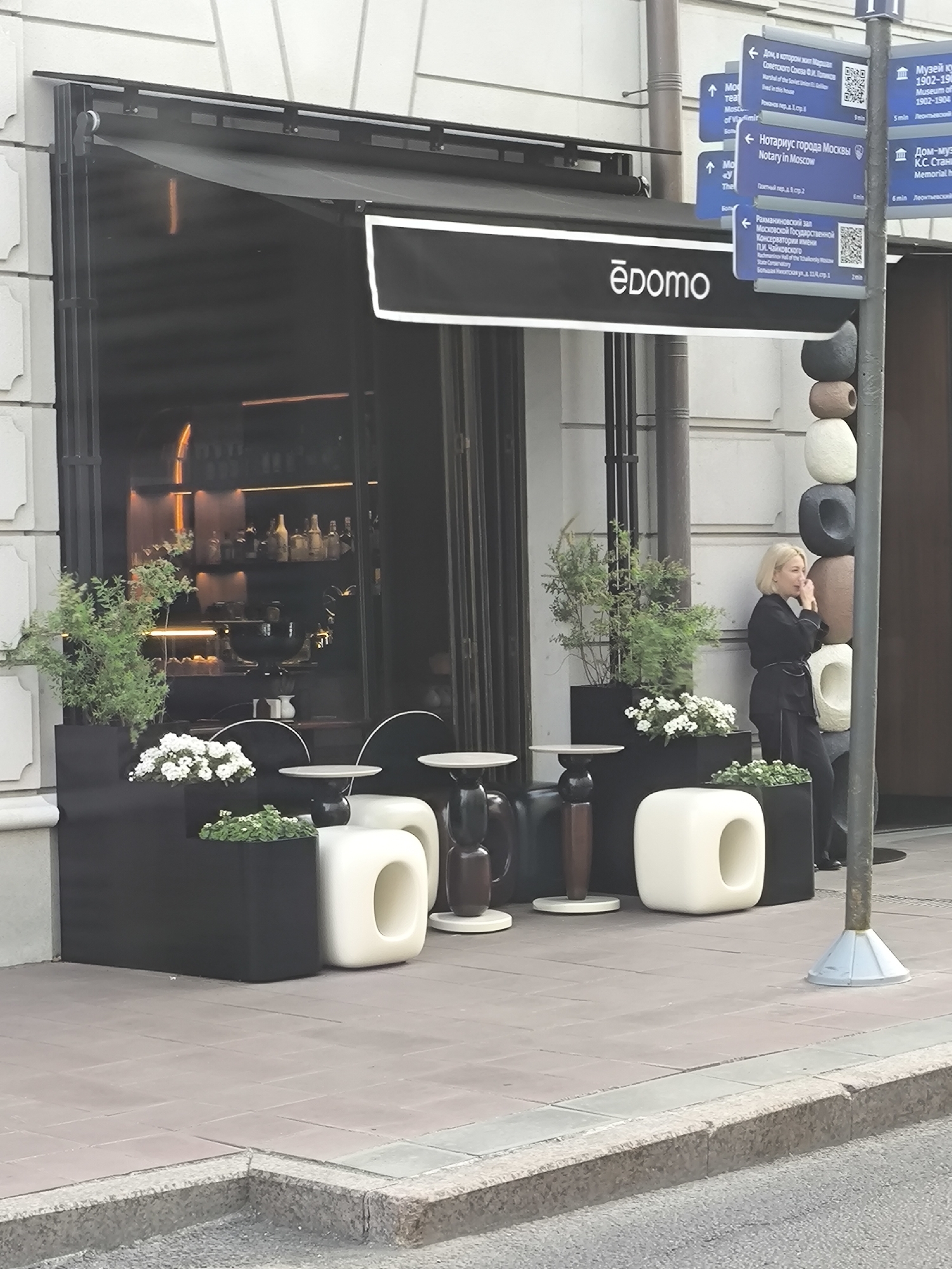 Here is a description of the image:
The image shows the exterior of a business, possibly a restaurant or café, named "ēdomo." It features a black awning with the business name written in white. The entrance area has outdoor seating with modern, minimalist black and white furniture, including planters with flowers and greenery. A blonde woman in dark clothing is standing near the entrance, appearing to be on a phone call.
To the right of the entrance, there is a pillar made of stacked stone or ceramic pieces, and further to the right, a street sign with multiple blue directional signs pointing towards local attractions such as "Moscow Theatre," "Notary in Moscow", "Rachmaninoff Hall" and "Museum of Moscow." Some of the signs include QR codes.
The location appears to be urban. The street and sidewalk are visible in the foreground.