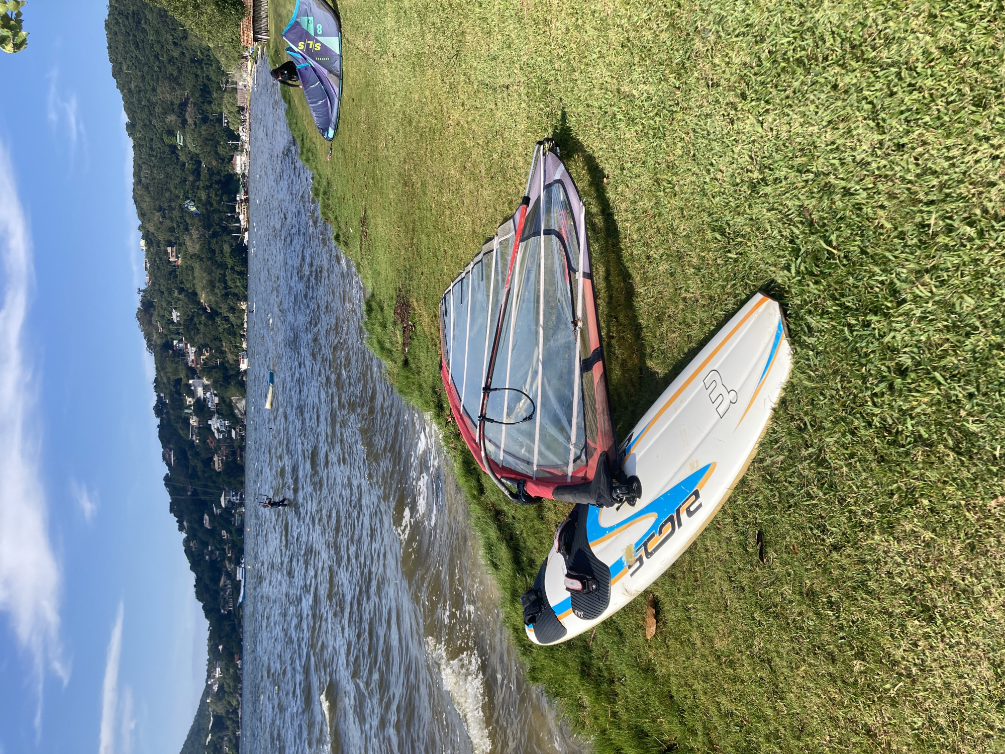The image shows a windsurfing setup on the grassy bank of Lagoa da Conceição in Florianópolis, Brazil. A white windsurfing board with blue and orange accents, labeled "STAR" and "R", lies next to a windsurfing sail with a red frame. In the background, the lake stretches out, with a densely forested hillside and buildings visible on the far shore. Two individuals appear to be kitesurfing in the distance on the lake. The sky is clear and blue. Another windsurfing setup is visible further away, on the bank.