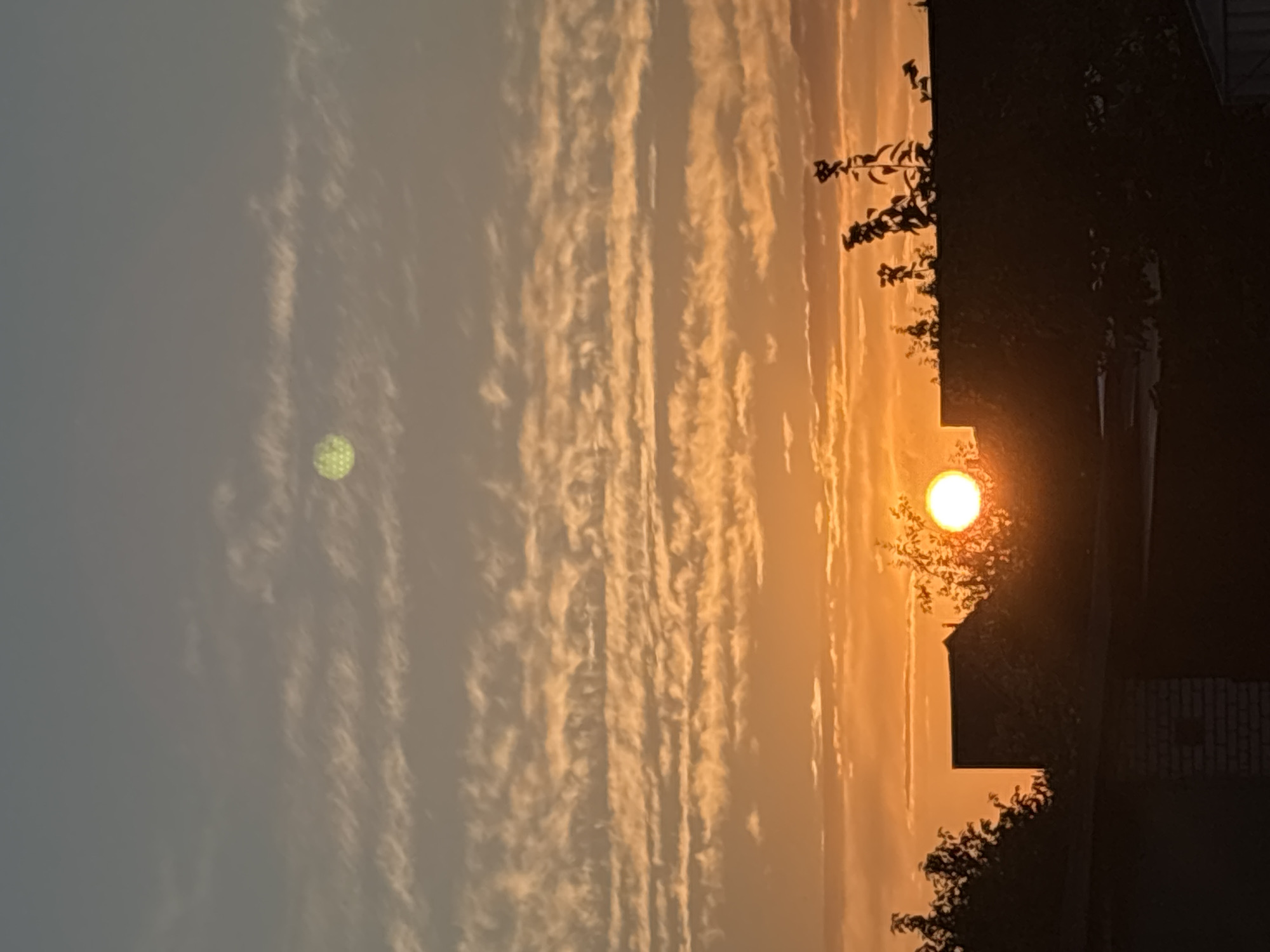 The image shows a sunset over a residential area. The sky is filled with layers of clouds, with the lower ones glowing orange from the setting sun and the upper ones a pale gray. The sun itself is a bright orb, partially obscured by trees and the rooftops of buildings. In the upper portion of the sky, a smaller, circular object with a textured, greenish appearance is visible, possibly a lens flare or another atmospheric phenomenon. The foreground is dominated by the dark silhouettes of trees and houses, suggesting a suburban or rural setting. The overall scene evokes a sense of calm and natural beauty. Based on the provided context, this scene is located in the city of Kapyĺ, Belarus.