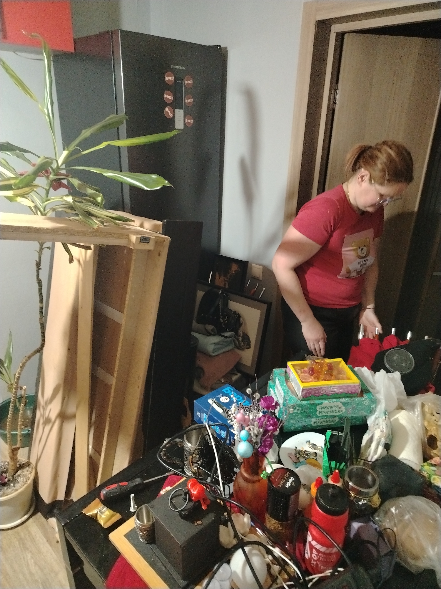 The image depicts an indoor scene in what appears to be a home in Chaikovsky, Russia, characterized by a cluttered environment.
A woman with auburn hair, tied in a ponytail and wearing glasses, is the primary human subject. She is dressed in a red t-shirt featuring a cartoon teddy bear graphic with partially visible text "DO IT MY...", and dark pants. She is positioned on the right side of the frame, looking down intently at a table, suggesting she is engaged in sorting or preparing items.
To the left, a tall houseplant with green and white variegated leaves stands in a pot on the floor. Behind it, a dark-colored Thomson refrigerator is visible, featuring circular buttons on its front panel, some labeled "SUPER FRIGOR" (partially visible). Adjacent to the plant and refrigerator, a large wooden frame or disassembled shelf leans against the wall. Further in the background, a light-colored wooden door stands slightly ajar.
The foreground is dominated by a black table overflowing with a diverse array of objects. These include a yellow box filled with what appears to be candies or jellies, resting on a green box with Cyrillic text. Other items on the table include a red and black screwdriver, metal cups, several bottles (one red with "TOP" and "POP" visible, another a "Jardin DEEP DARK" spice container), artificial flowers in a small vase, a white plate, and various plastic bags, some seemingly containing food. The scene suggests a moment of domestic activity, possibly tidying, unpacking, or preparing for an event.
