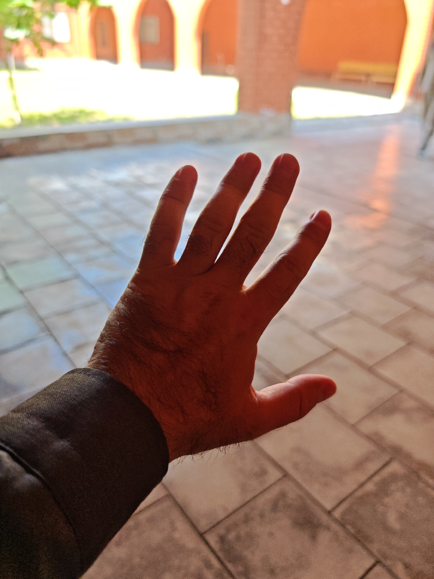 A person's hand, palm facing forward, is held up in front of a tiled walkway. The person is wearing a dark sleeve on their left arm. In the background, there are arched openings leading into a building with an orange-colored facade, suggesting an outdoor public space. The scene is lit by natural sunlight, casting shadows on the tiles. The location is identified as Mayrtup, Russia.