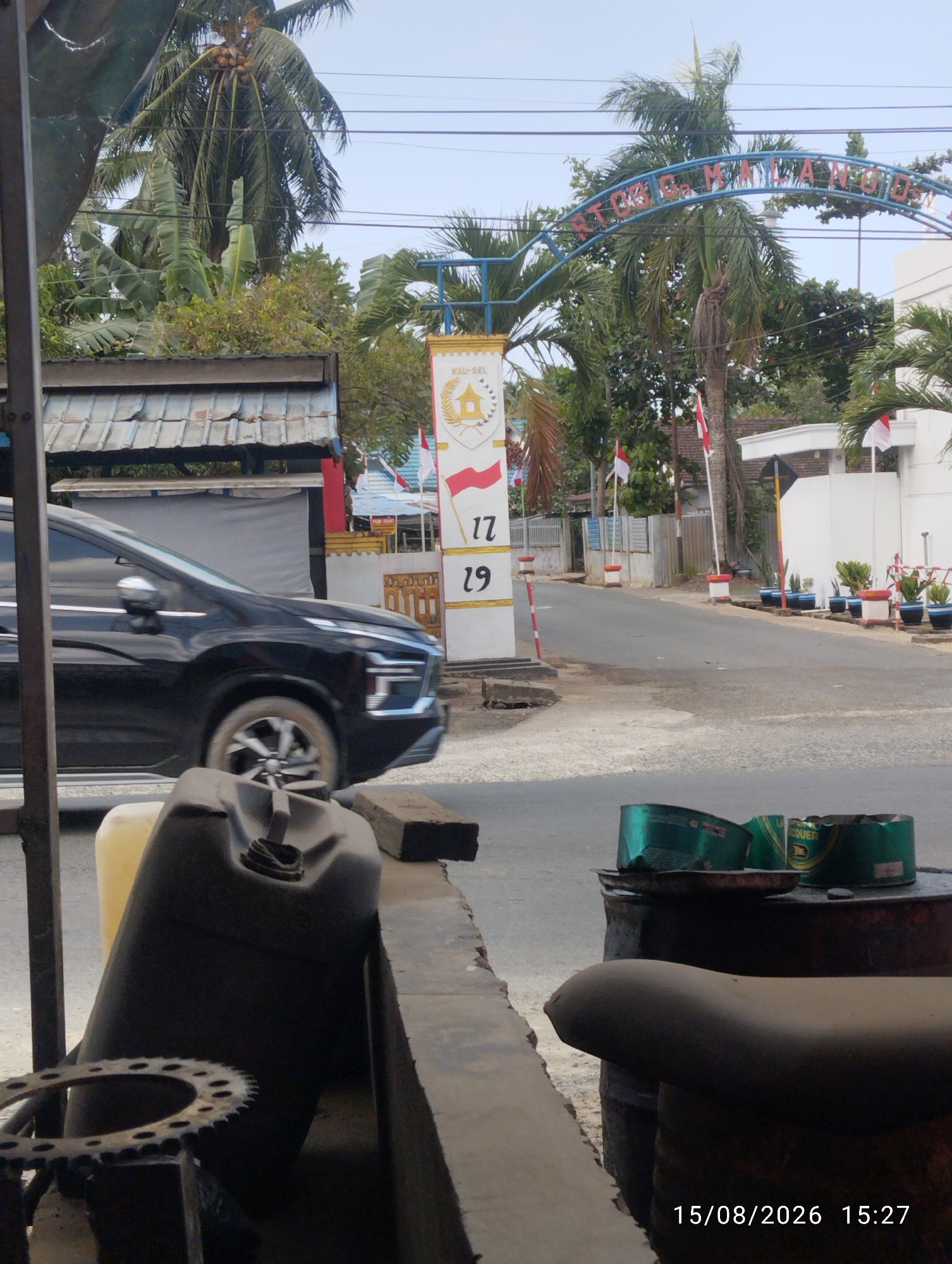 The image captures a street scene in Tegal Rejo, Indonesia. In the foreground, a black car is driving past. Partially visible in the foreground are several objects: a dark-colored jerrycan, a yellow container, and a metal sprocket. In the mid-ground, a white pillar with red and white flags and numbers "17" and "29" is prominent, likely a landmark or signpost. A street stretches out behind it, lined with trees and buildings. The background shows a blue archway with text and more flags. The overall environment appears to be an outdoor urban or semi-urban setting.