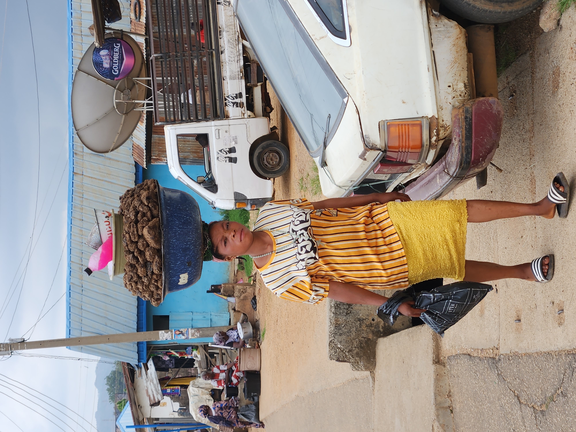 This image, rotated 90 degrees clockwise, depicts a vibrant street scene likely in Kabong, Nigeria, centered around a woman balancing a large load on her head.
The main subject is a woman standing on a concrete surface, facing the camera with a neutral expression. She wears a short-sleeved yellow and white striped top, a textured yellow skirt, and black-and-white striped flip-flops. On her head, she expertly balances a large, dark blue basin overflowing with numerous dark brown, irregularly shaped items, possibly yams, nuts, or prepared food. In her left hand, she carries a black plastic bag with the brand name "VIVA" visible on it.
In the background, to her right, is the rear of an older model white sedan showing signs of wear and rust on its bumper and bodywork. Behind that is a white pickup truck with a metal cage structure on its bed. To the left, several buildings with light blue corrugated metal roofs are visible, and a large grey satellite dish with a circular sign displaying "GOLDBERG" (a Nigerian beer brand) is mounted on one roof. Further into the background, other individuals are seated or standing near what appear to be market stalls. A utility pole and overhead wires run along the left side of the frame. The sky appears bright but somewhat hazy, suggesting daytime. The overall setting is an active, unpaved or partially paved street or market area, bustling with daily commerce.