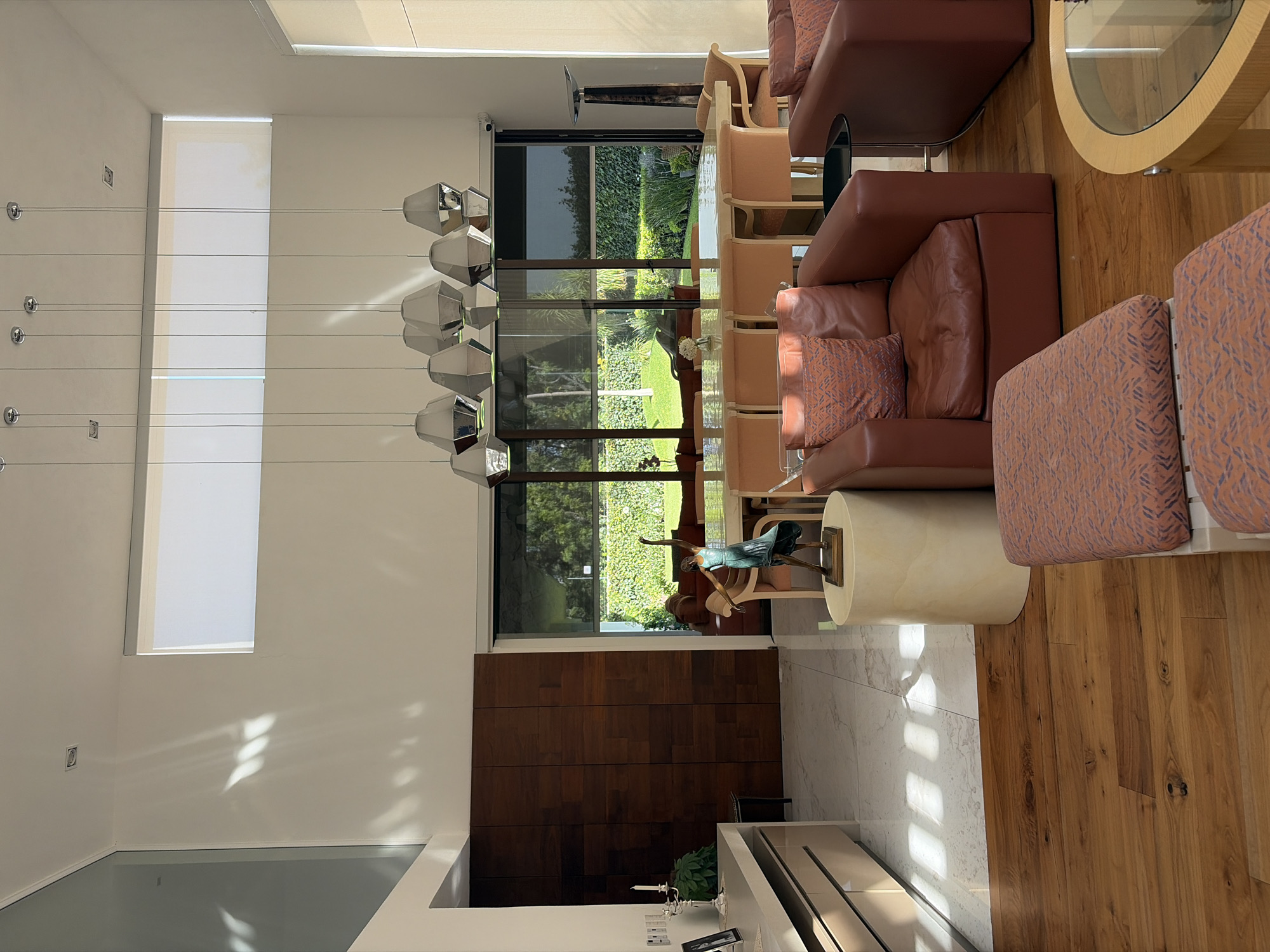 This image captures a brightly lit, modern living and dining area of a residence, likely located in San Bernardino Tlaxcalancingo, Mexico. The space features an open-plan layout with large windows overlooking a lush green garden.
The living area is furnished with comfortable brown leather sofas adorned with patterned cushions in shades of orange, peach, and blue. A round wooden coffee table with a glass top and a cylindrical cream-colored side table accompany the seating arrangement. A unique green sculpture of a dancing figure is placed on the side table. Adjacent to this, a dining area is visible, featuring a long light wood table with a clear glass top, surrounded by light wood dining chairs. A modern chandelier with multiple geometric, metallic light fixtures hangs above.
Architectural details include tall white walls, one of which is accented with rich dark wood paneling. The flooring transitions from light wood in the living area to light-colored marble or tile closer to a white console table. Sunlight streams through the large windows, casting distinct patterns of light and shadow across the floors and walls, indicating a sunny day, likely mid-morning or afternoon.
The exterior view through the expansive windows reveals a well-maintained garden with various green foliage, trees, and bushes, creating a tranquil backdrop. No people are visible in the image, suggesting a serene and unoccupied space. There is no visible text.