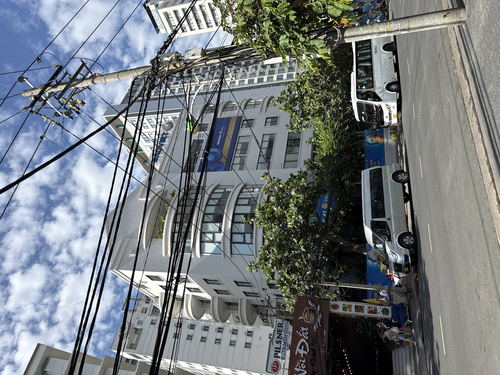 The image depicts a street scene in Nha Trang, Vietnam, characterized by modern architecture and bustling urban activity. On the right side of the image, a concrete utility pole, heavily laden with black electrical wires, stands prominently. Behind the pole, a white multi-story building displays signage that appears to be in Vietnamese. To the left of the pole, two white vehicles are parked along the roadside: a smaller van and a larger bus.
The central focus of the image is a row of contemporary white buildings. The tallest building on the left has a repeating pattern of windows. Adjacent to it, a building with a curved facade features multiple balconies and large windows. Above the entrance to a establishment at street level, a prominent sign displays "33 Pilsner EXTRA SMOOTH," suggesting a restaurant or bar. Further down the street, another building exhibits a large blue banner with text and images, possibly an advertisement.
In the foreground, the asphalt street is marked with yellow lines. On the sidewalk to the left, two women are walking, one wearing a hat and the other a striped shirt and shorts. Another person, dressed in shorts and a light-colored shirt, stands near the vehicles. Several trees with green foliage are interspersed throughout the scene, providing a touch of nature amidst the urban landscape. The sky above is a bright blue, dotted with fluffy white clouds, indicating fair weather. The overall impression is that of a lively street in a Vietnamese city, with a mix of commercial establishments and residential or office buildings.