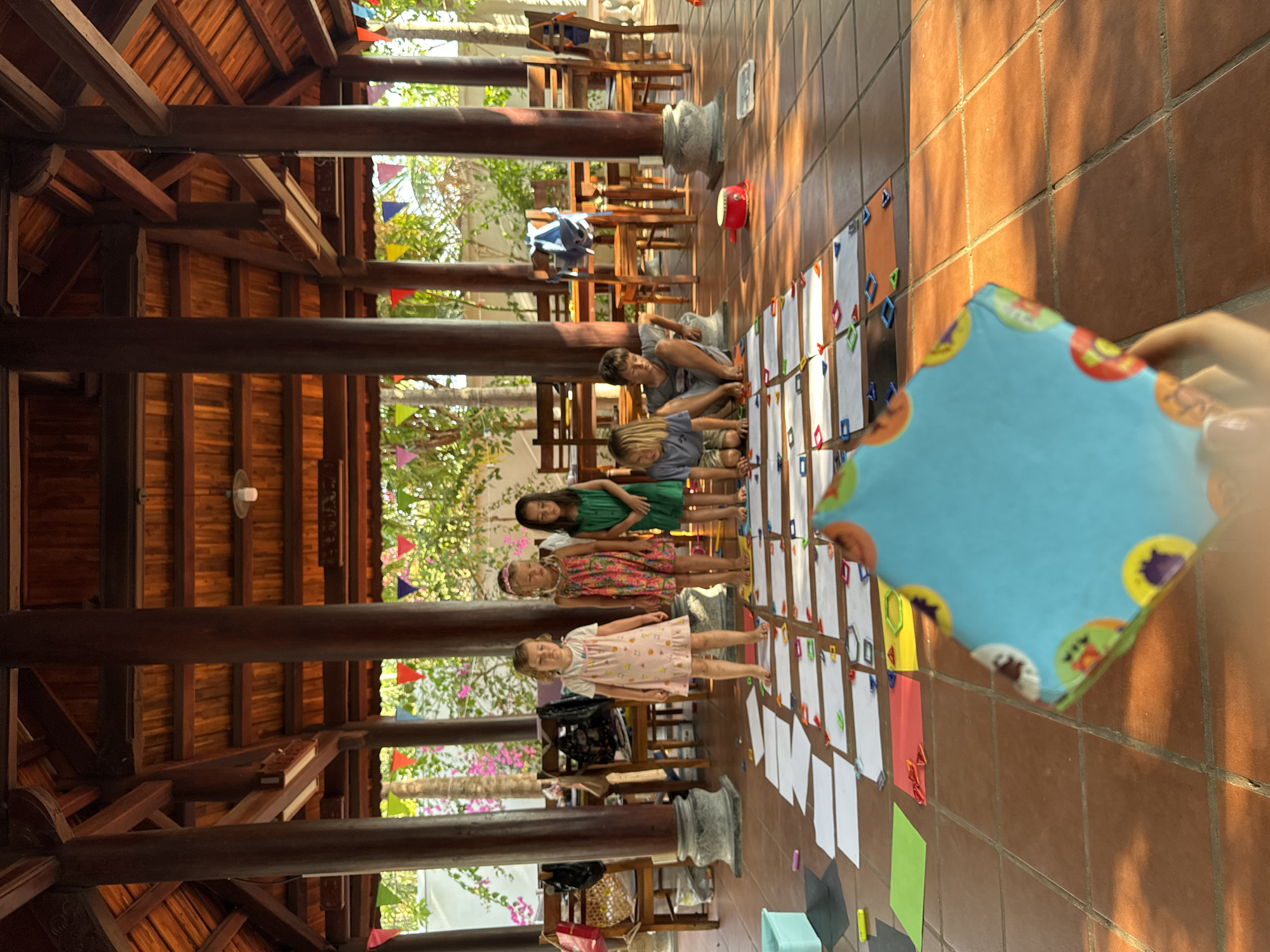 The image captures a scene in Mũi Né, Vietnam, under a wooden-roofed structure supported by wooden pillars. Four children stand on a line of papers taped to the tiled floor, each paper adorned with colorful shapes. A man is sitting on the floor, presumably interacting with the papers or the children. A blue, square object with colorful circles around the edges is in the extreme foreground, obscuring part of the view. In the background, wooden chairs and tables are arranged under the structure. Exterior vegetation, including flowering trees and colorful flags, is visible beyond the roof.