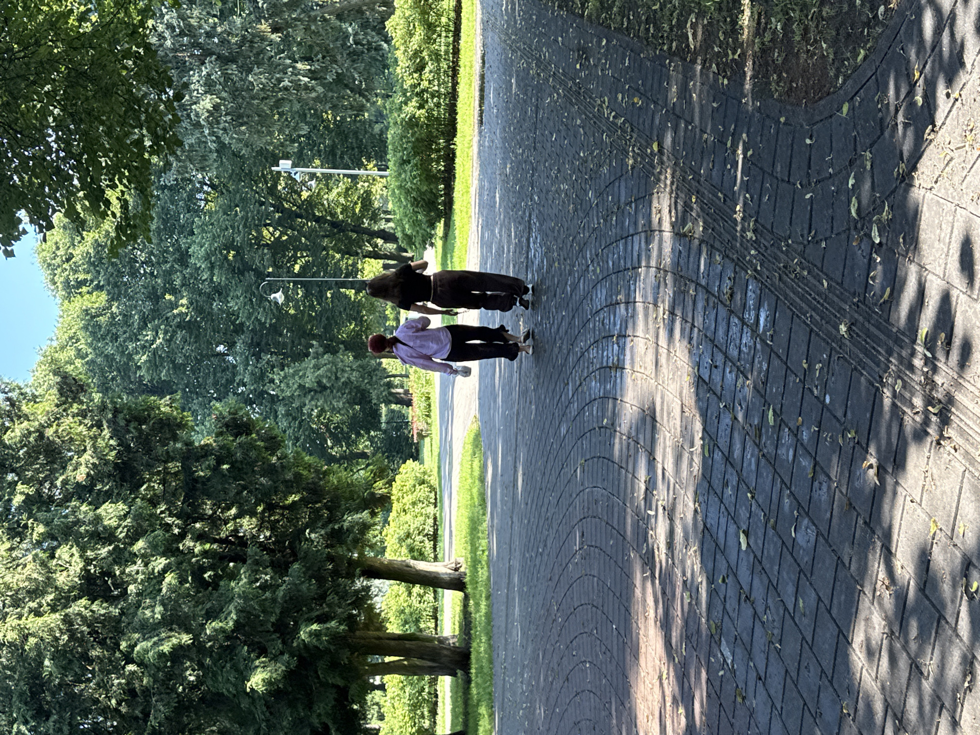 The image shows two individuals walking away from the camera on a paved pathway in a park, likely located in Riga, Latvia, given the characteristic architecture and lush greenery common in the city's parks. The path is made of dark, rectangular pavers arranged in a circular pattern, with scattered fallen leaves. To the left of the path, several trees with dense green foliage are visible, casting dappled shadows on the ground. In the background, more trees and manicured bushes form a dense green backdrop under a clear blue sky. Two lampposts are present in the mid-ground, one with a curved design and the other a more standard urban lamppost. The individuals are dressed casually; one is wearing a light pink long-sleeved shirt and dark pants, holding a white object in their right hand. The other is wearing a black crop top and dark brown sweatpants. The lighting suggests it is daytime, likely late morning or early afternoon, with the sun creating strong contrasts of light and shadow. The overall impression is a serene outdoor scene in a well-maintained park.