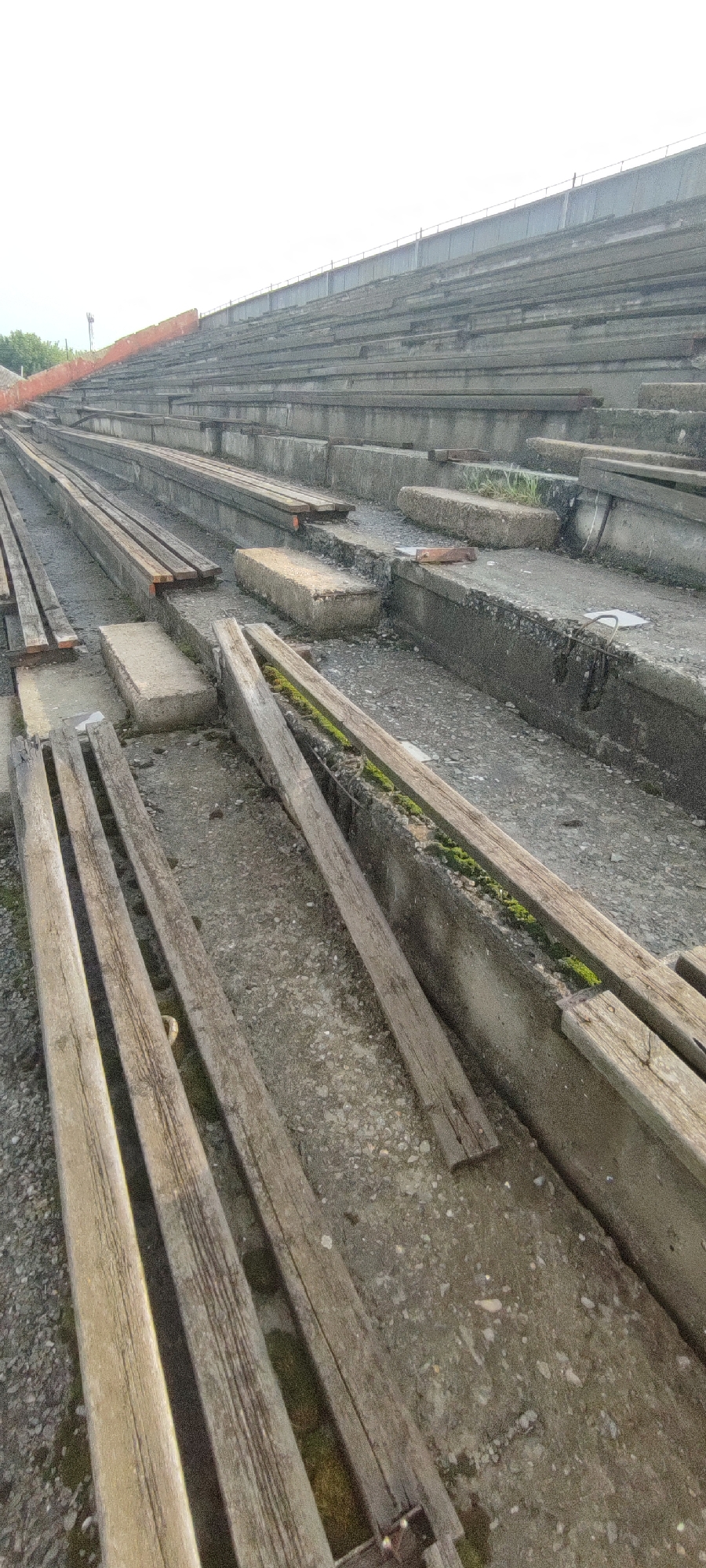 The image shows a section of an abandoned outdoor stadium with concrete tiered seating. Several rows of concrete bleachers are visible, some with wooden planks that appear to be remnants of seats. The area is weathered and overgrown with patches of moss and grass. The sky is overcast and bright. The overall impression is one of disuse and decay.