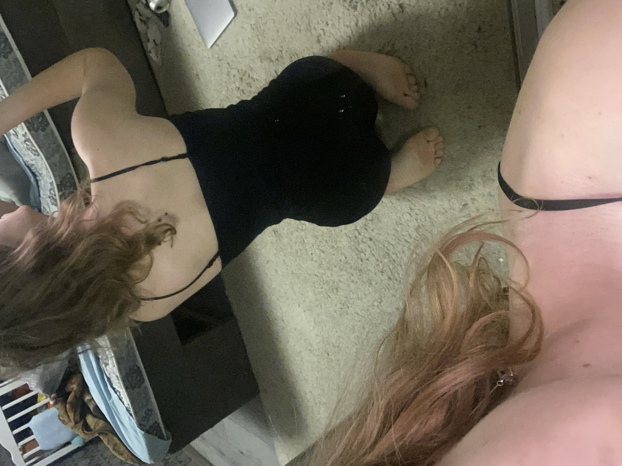 The image shows two individuals in an indoor setting. One is lying on a carpet, wearing a black dress. The other individual is close to the camera, with only their upper body visible, including a bare shoulder and chest area with a thin black strap, likely from underwear or a top. Their hair is reddish-blonde and partially obscures their face. The background includes a carpet, part of a bed or couch with patterned fabric, and a white shelf-like object. There's no visible indication to suggest the location is Ternopil, Ukraine.