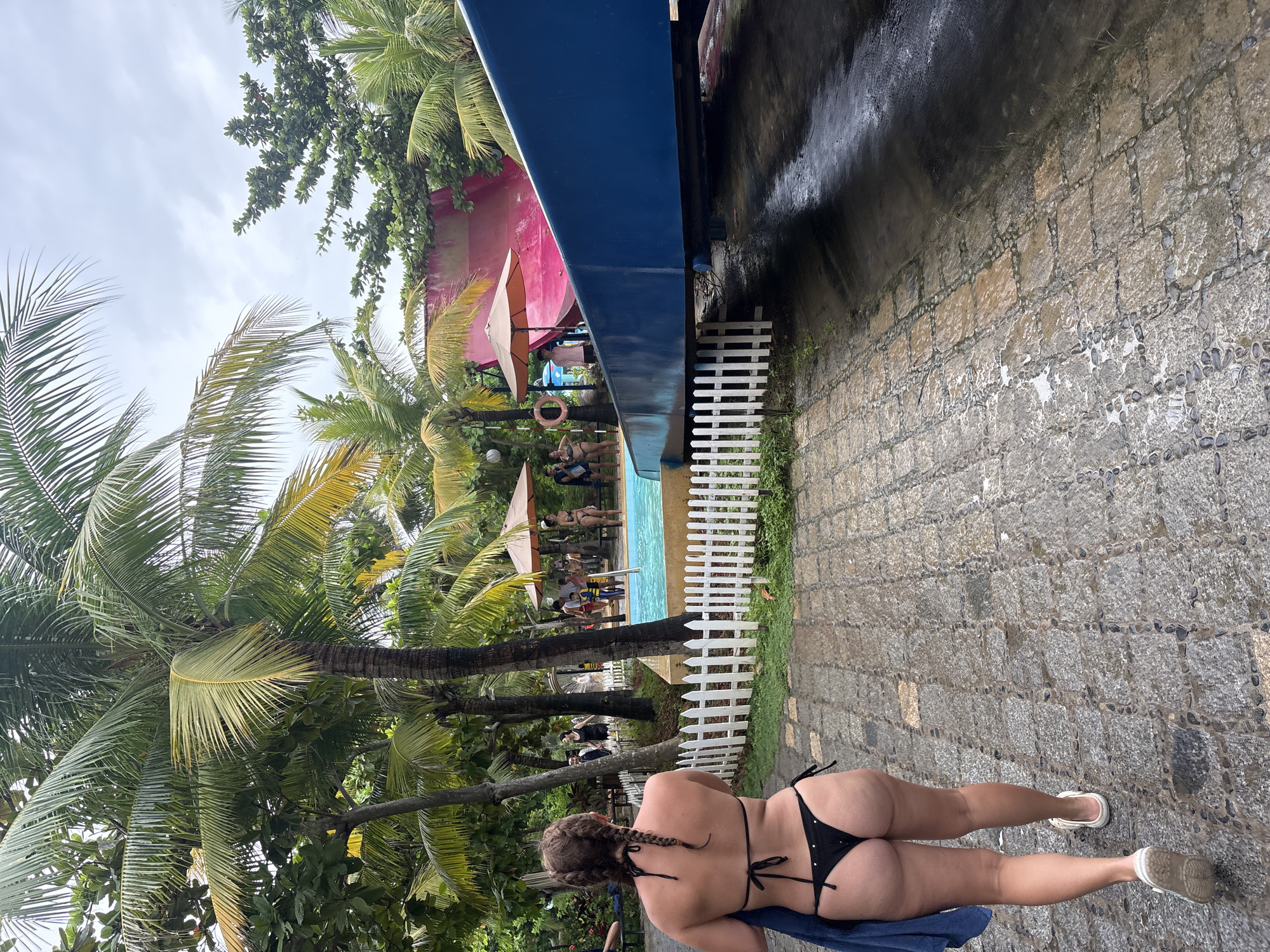 A woman in a black bikini walks away from the camera on a wet stone path, carrying a blue towel. She has braided hair. In the background, palm trees line a pool area with a pink water slide and several people, some of whom are standing by the pool or near umbrellas. The sky is overcast. The scene appears to be at a water park in Nha Trang, Vietnam.