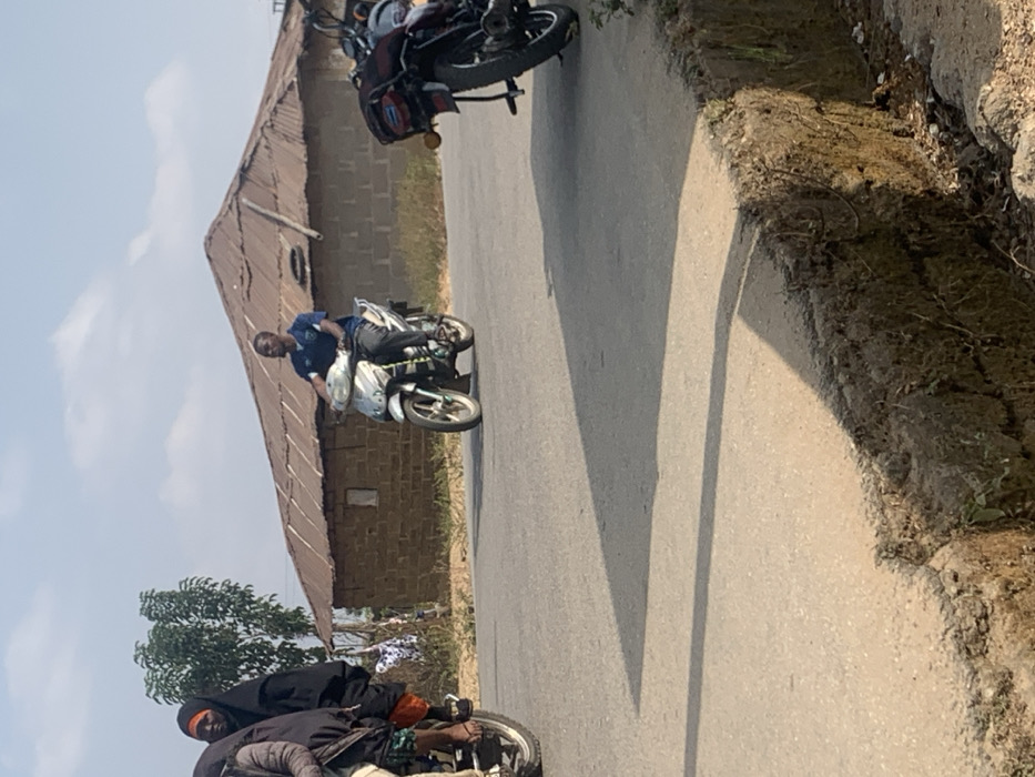 The image captures an outdoor street scene in Nagazi, Nigeria, under a clear sky with visible long shadows, indicating a sunny mid-morning or late afternoon. Three individuals are present on motorcycles. In the mid-ground, a male wearing a dark blue shirt and dark trousers rides a silver scooter-style motorcycle, looking towards the left. To the left foreground, the partial view of a dark motorcycle shows two occupants: a primary rider (partially obscured) and a passenger in a black hijab over an orange garment. On the right foreground, a dark motorcycle with a luggage rack is partially visible, oriented away from the viewer, appearing stationary. The road is paved with asphalt, showing a significant eroded dirt embankment and ditch on its right side. In the background, a low-rise building constructed of light-brown concrete blocks features a weathered, reddish-brown corrugated metal roof with visible wooden supports. Sparse vegetation, including trees and dry foliage, is present in the background and along the road's edges.
