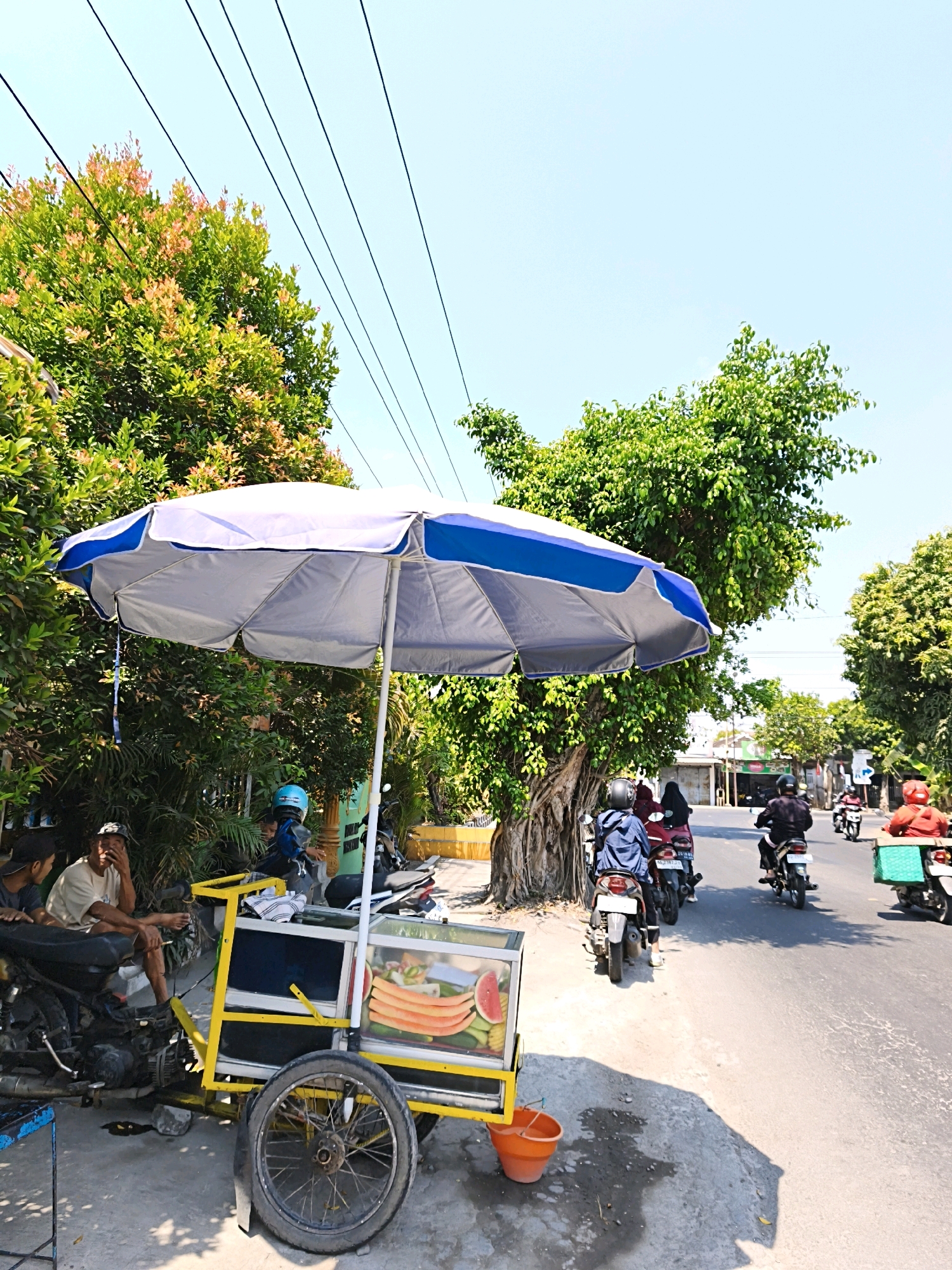 In Desa Sawahan, Indonesia, on a sun-drenched street, a fruit cart stands prominently under a large blue and grey umbrella. The cart is laden with various sliced fruits, including watermelon and other colorful produce, displayed in a clear glass enclosure. Beside the cart, a man in a light-colored t-shirt and cap sits on a motorcycle, his hand near his face, appearing to be in conversation with another person partially visible to his left. Behind them, a young person wearing a blue helmet is seated on another motorcycle.
The street is active with motorcycle traffic. Several individuals on motorcycles are seen riding away from the viewer. One motorcycle in the foreground on the right has a green bin attached to its rear. Lush green trees and foliage line the left side of the street, providing shade in some areas, while buildings and shops are visible in the background on the right. The sky is clear and bright blue.