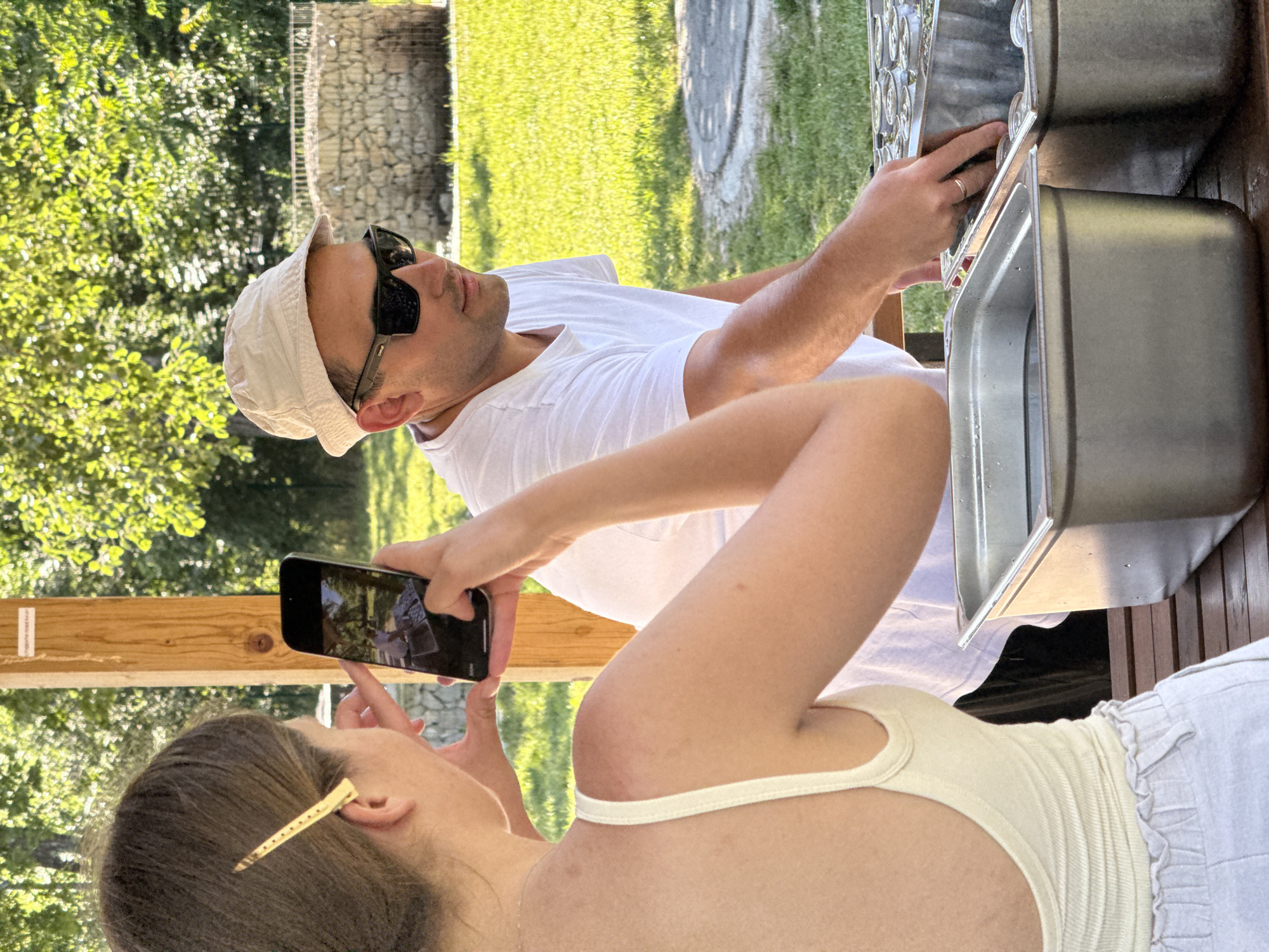 A woman takes a picture with her smartphone of a man who is placing a rack of glasses into a stainless steel sink. The scene takes place outdoors, with trees and grass visible in the background. The man is wearing a white t-shirt, a white bucket hat, and sunglasses. The woman is wearing a white tank top and has a hair clip. The location is Cukarica Urban Municipality, Serbia.